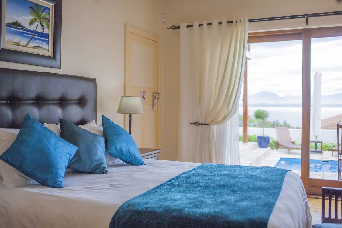Une chambre à coucher avec un grand lit, des oreillers bleus, une tête de lit, une table de chevet, une lampe et une vue sur l’extérieur grâce à une porte coulissante.