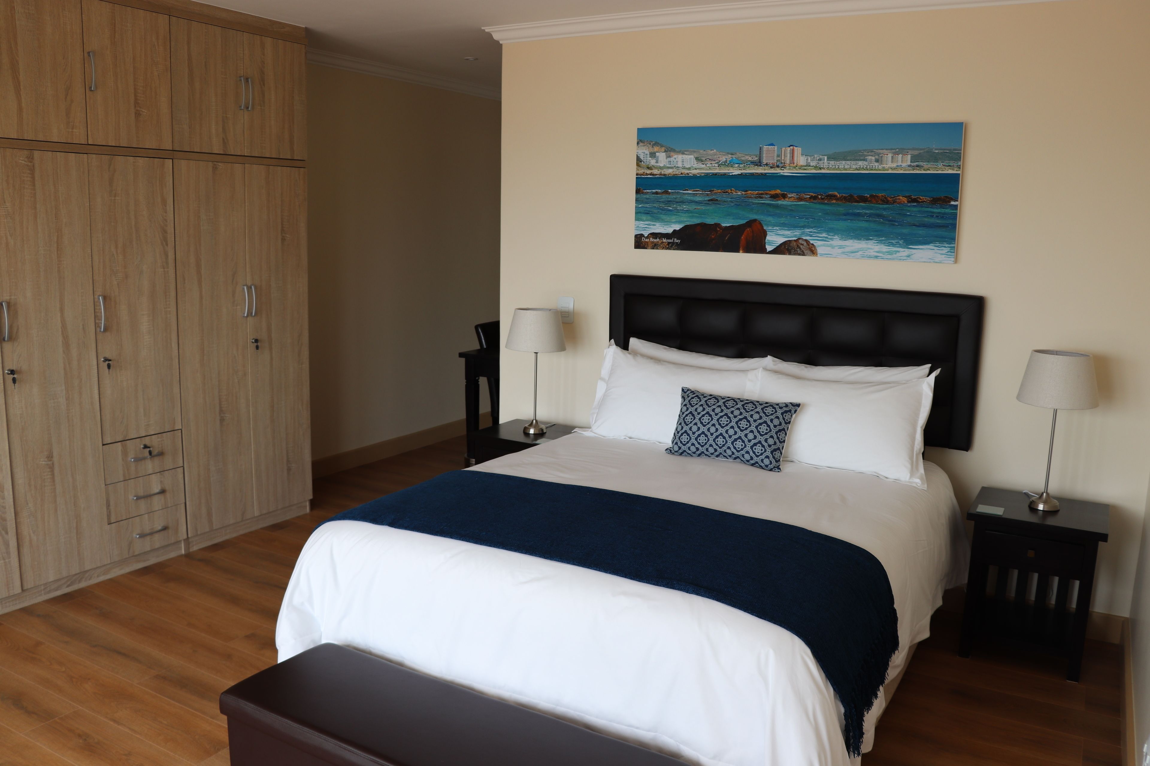 A bedroom with a large bed, two bedside tables with lamps, a wooden wardrobe, and a cityscape painting on the wall.