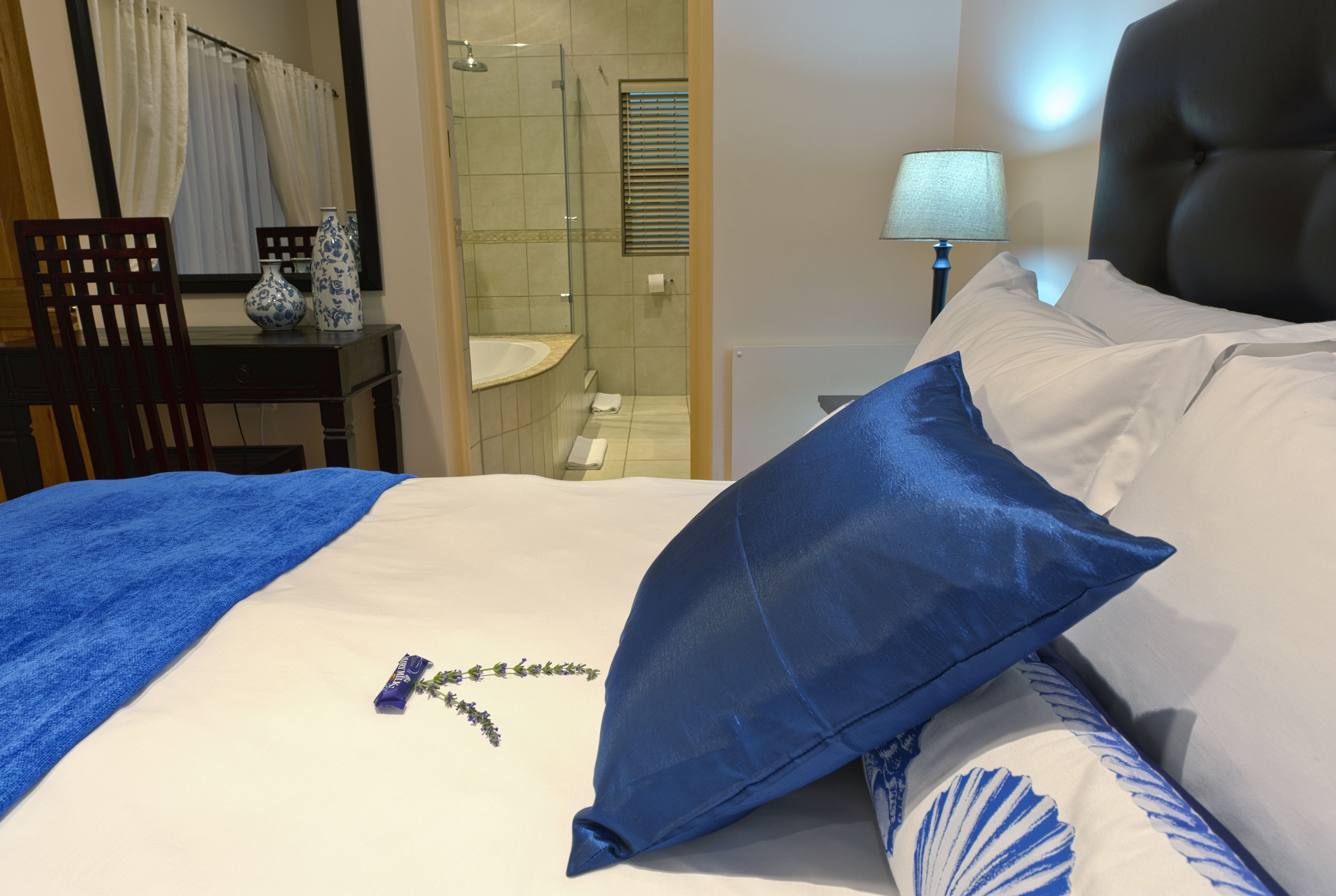 A hotel room with a bed, a nightstand with a lamp, a bathroom with a shower, and a desk with a chair.
