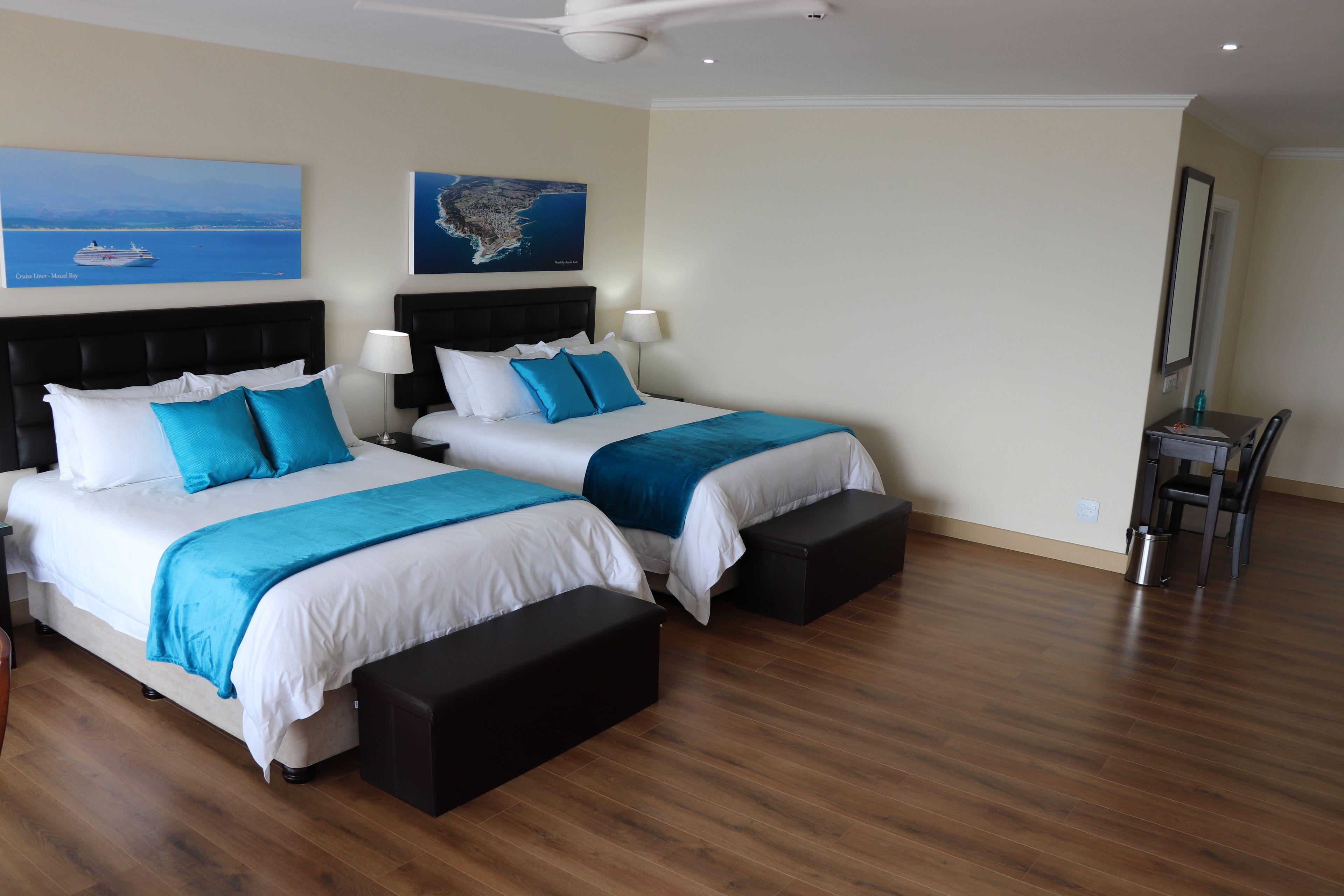 Habitación de hotel con dos camas, suelo de madera, ventilador de techo y cuadros en las paredes.