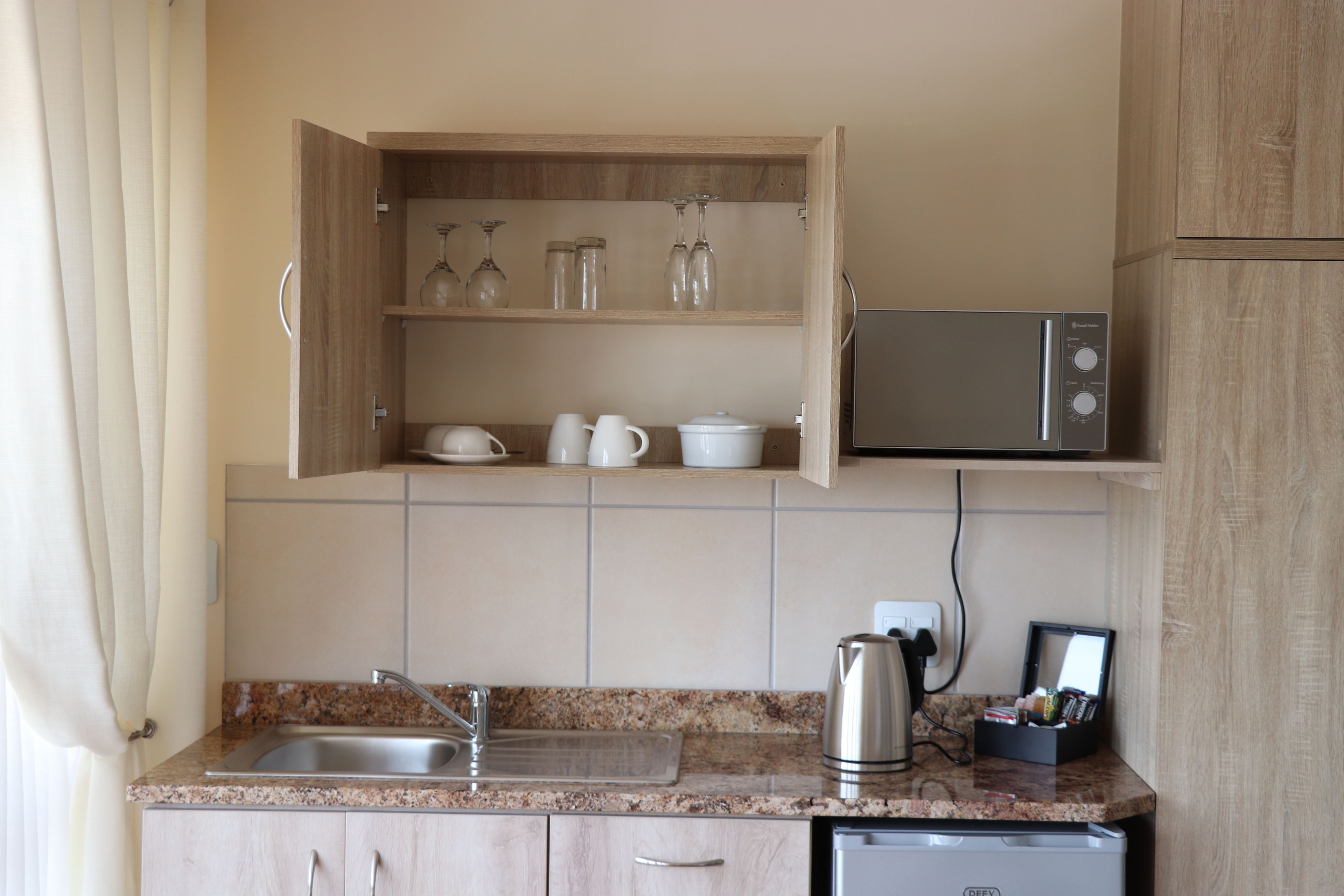 A kitchen with a granite countertop, stainless steel sink, microwave, and a cabinet with glassware and dishes.