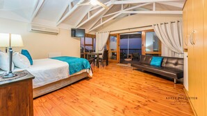 Une pièce spacieuse avec un plancher en bois, un lit recouvert d’une couverture bleue, un canapé avec un coussin bleu, une table à manger et un téléviseur.