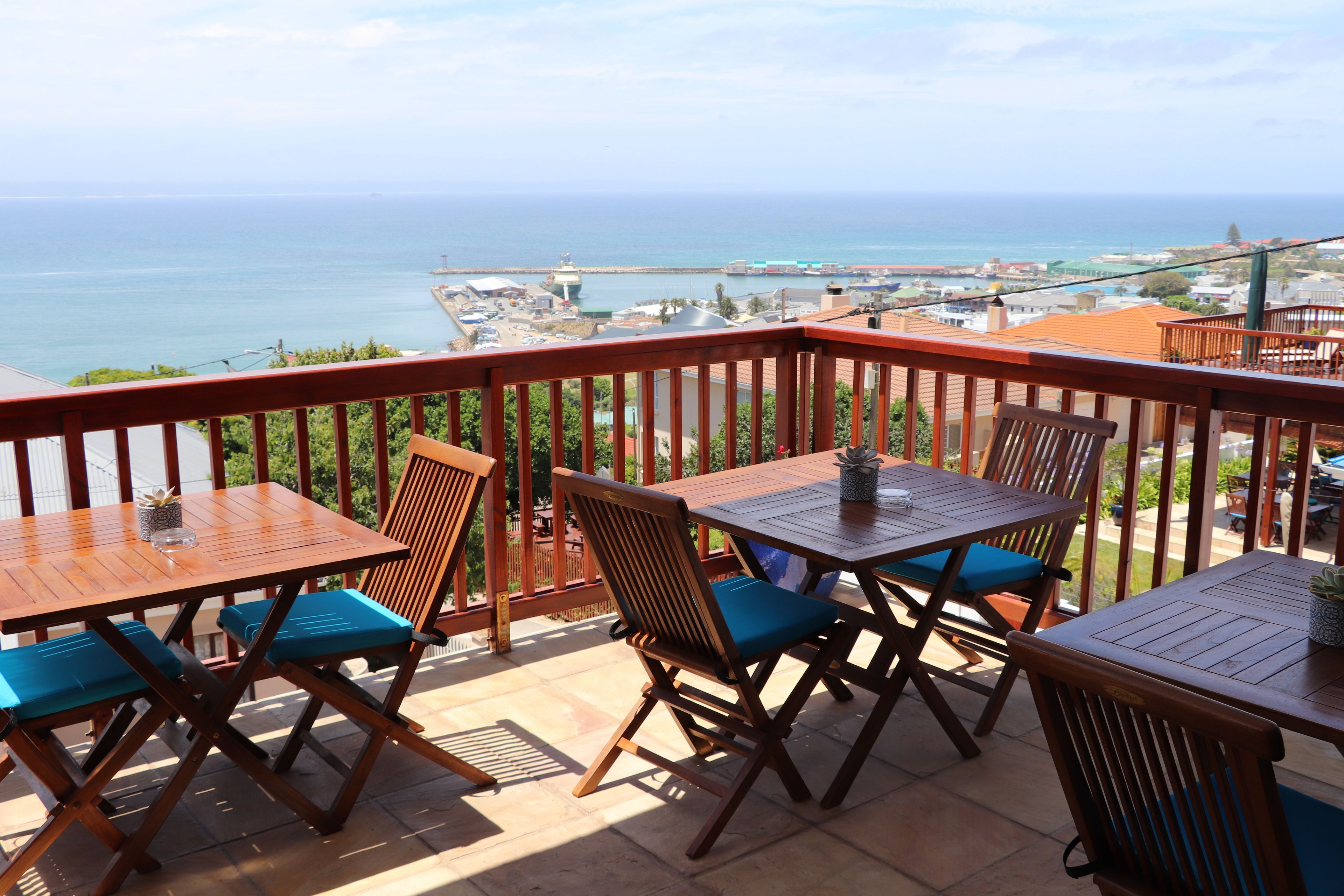 A balcony with wooden furniture, a view of the sea, and a distant cityscape.