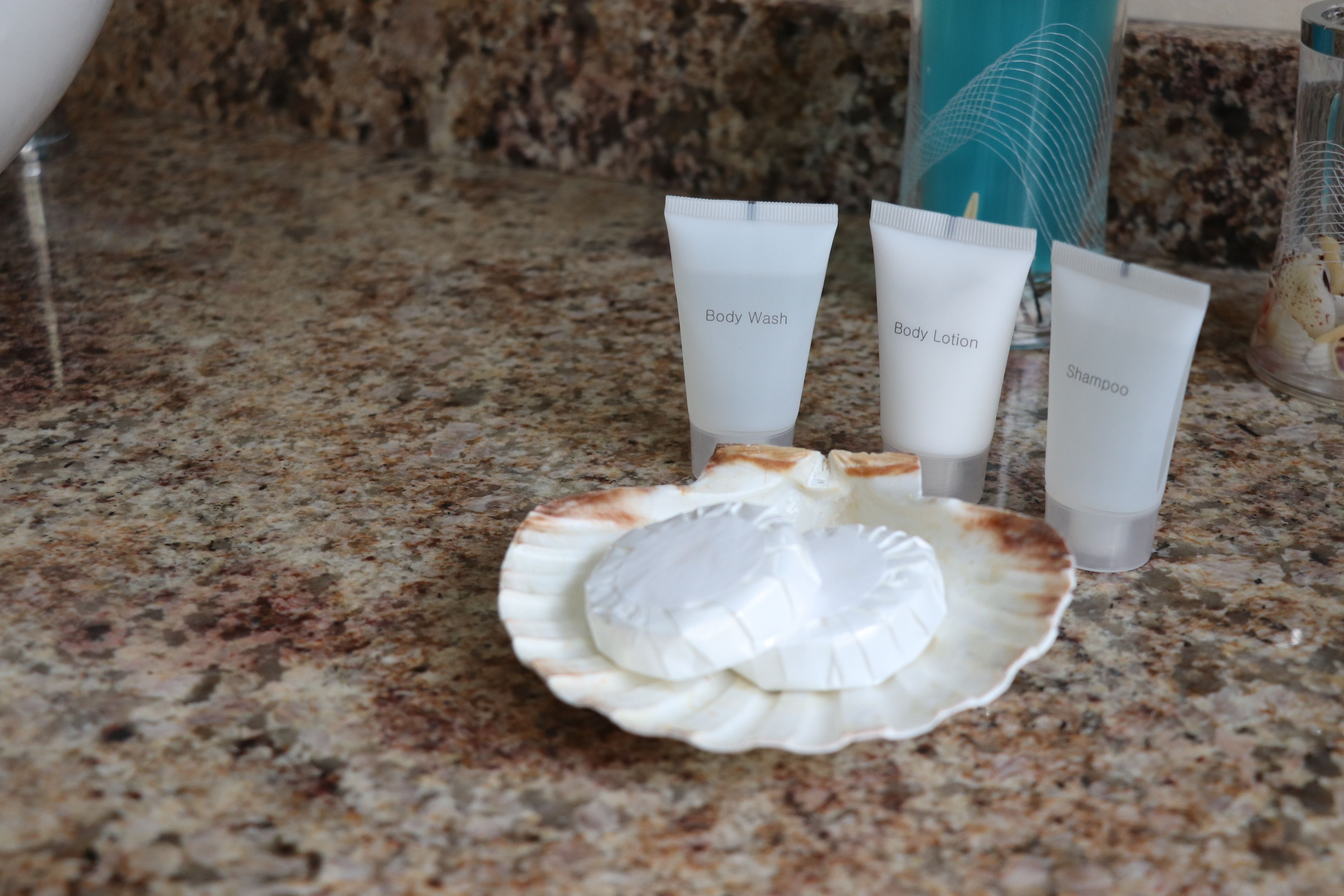 Un plan de travail en granit sur lequel reposent trois tubes de produits de toilette étiquetés « gel douche », « lotion pour le corps » et « shampoing », ainsi qu’un récipient en forme de coquillage.