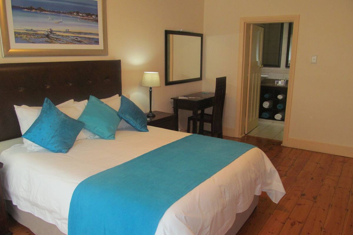 Une chambre d’hôtel avec un lit, un bureau, une chaise, une lampe et une salle de bain visible à travers une porte ouverte.