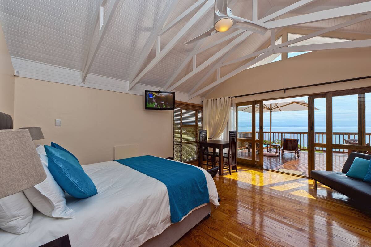 Une chambre avec un lit, une télévision, un balcon avec une table et des chaises, et une vue sur la mer.
