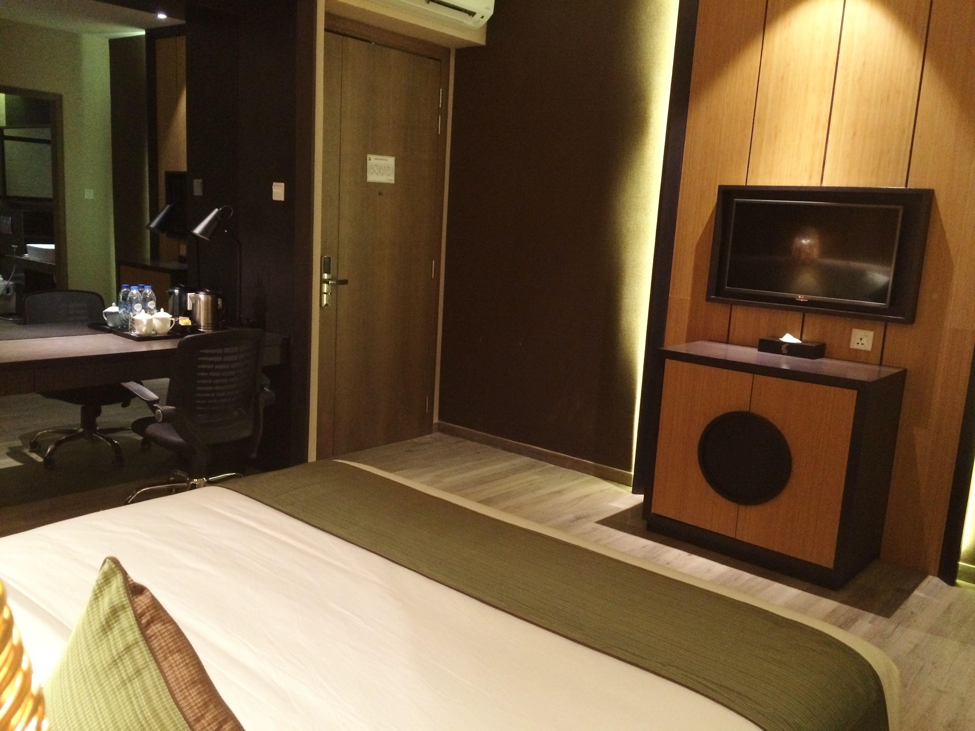 Una habitación de hotel con cama, escritorio con material de oficina, silla, televisor y un mueble pequeño.