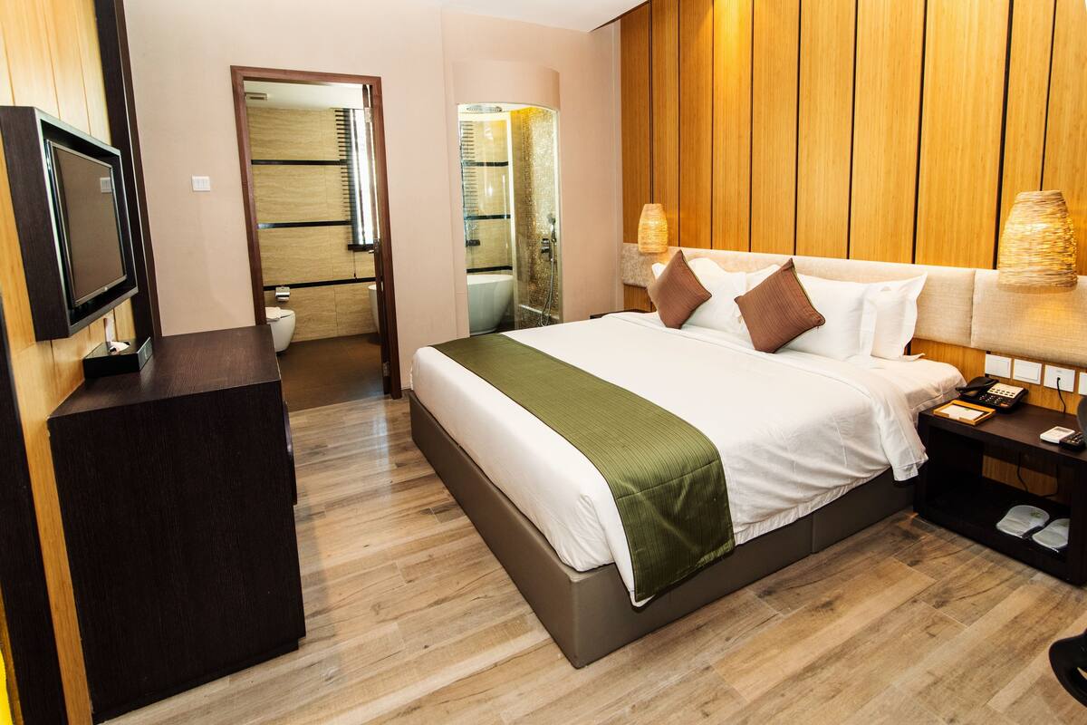 A hotel room with a large bed, a TV on a stand, a bedside table with a lamp, and a bathroom visible through an open door.