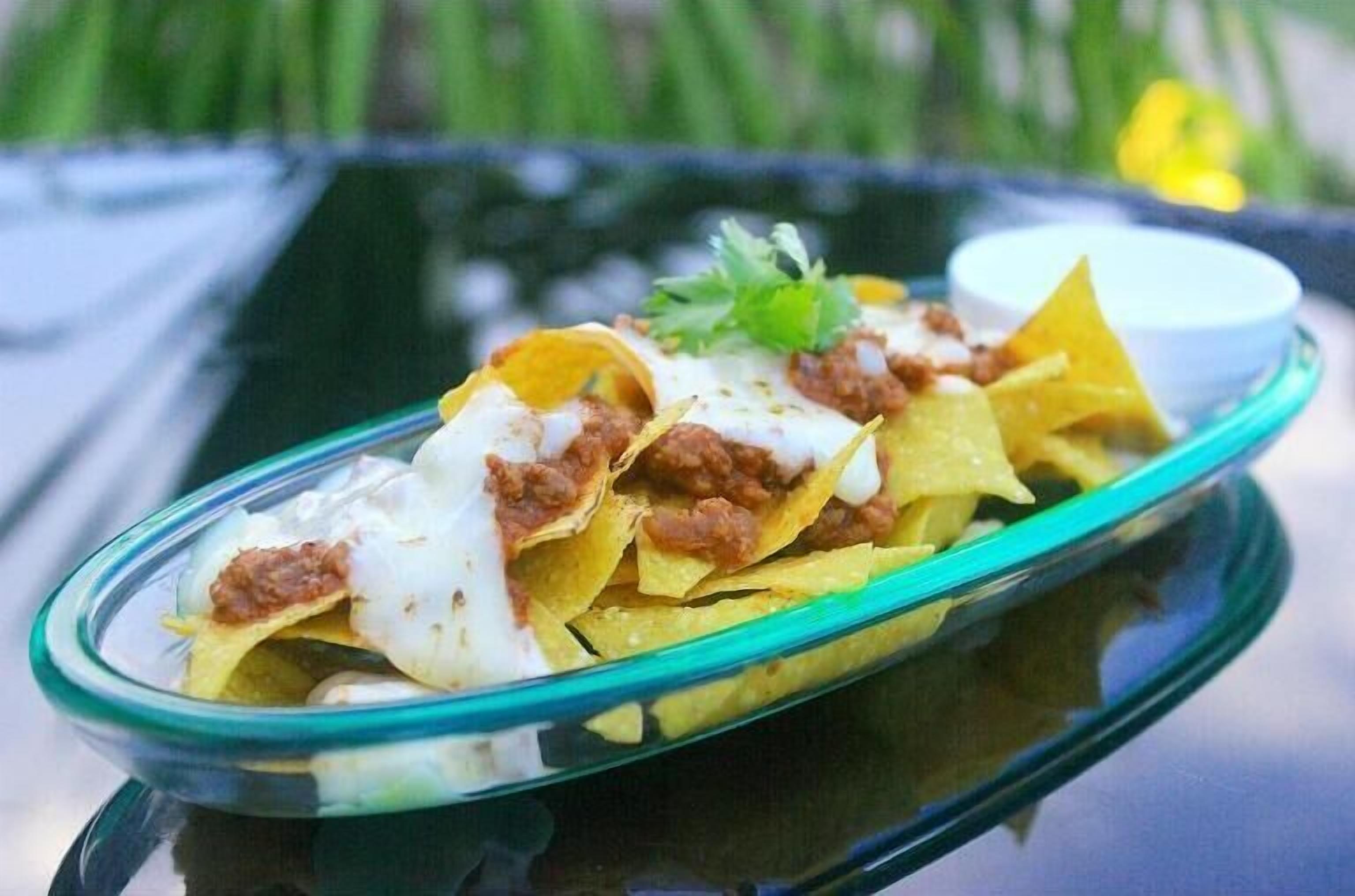 A glass dish with nachos topped with cheese and ground meat, accompanied by a small white bowl of sauce, on a reflective surface.