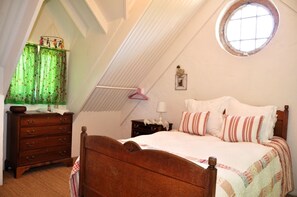 Une chambre avec un plafond en pente, une fenêtre ronde, un lit en bois, une commode et une table de chevet.