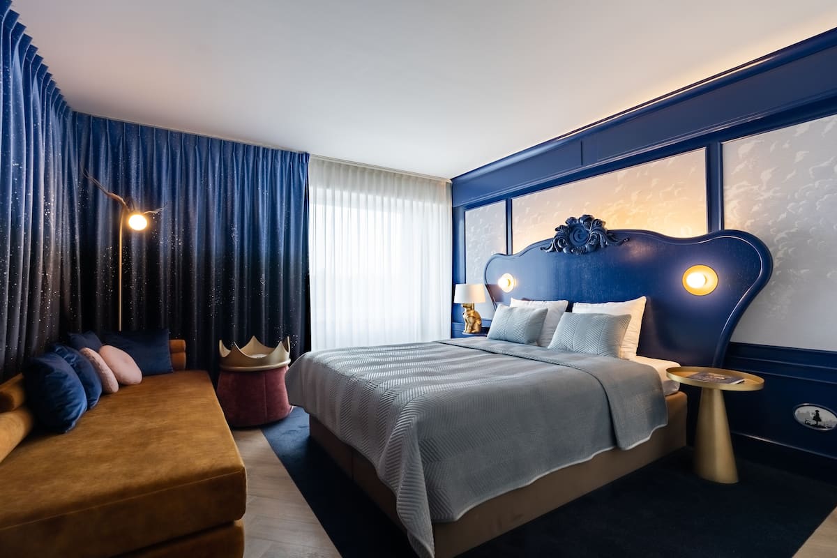 Quarto de hotel moderno com uma cama grande, uma mesinha lateral redonda dourada e uma parede de destaque azul escura.