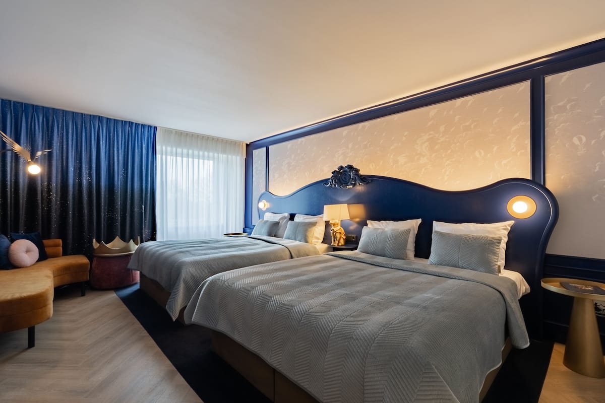 Quarto de hotel com duas camas, um sofá, uma mesa de cabeceira e uma luminária.