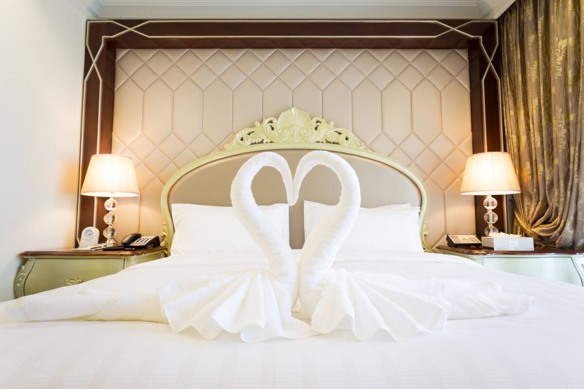 A hotel room with a bed featuring a heart-shaped towel arrangement, a tufted headboard, and two bedside tables with lamps and phones.
