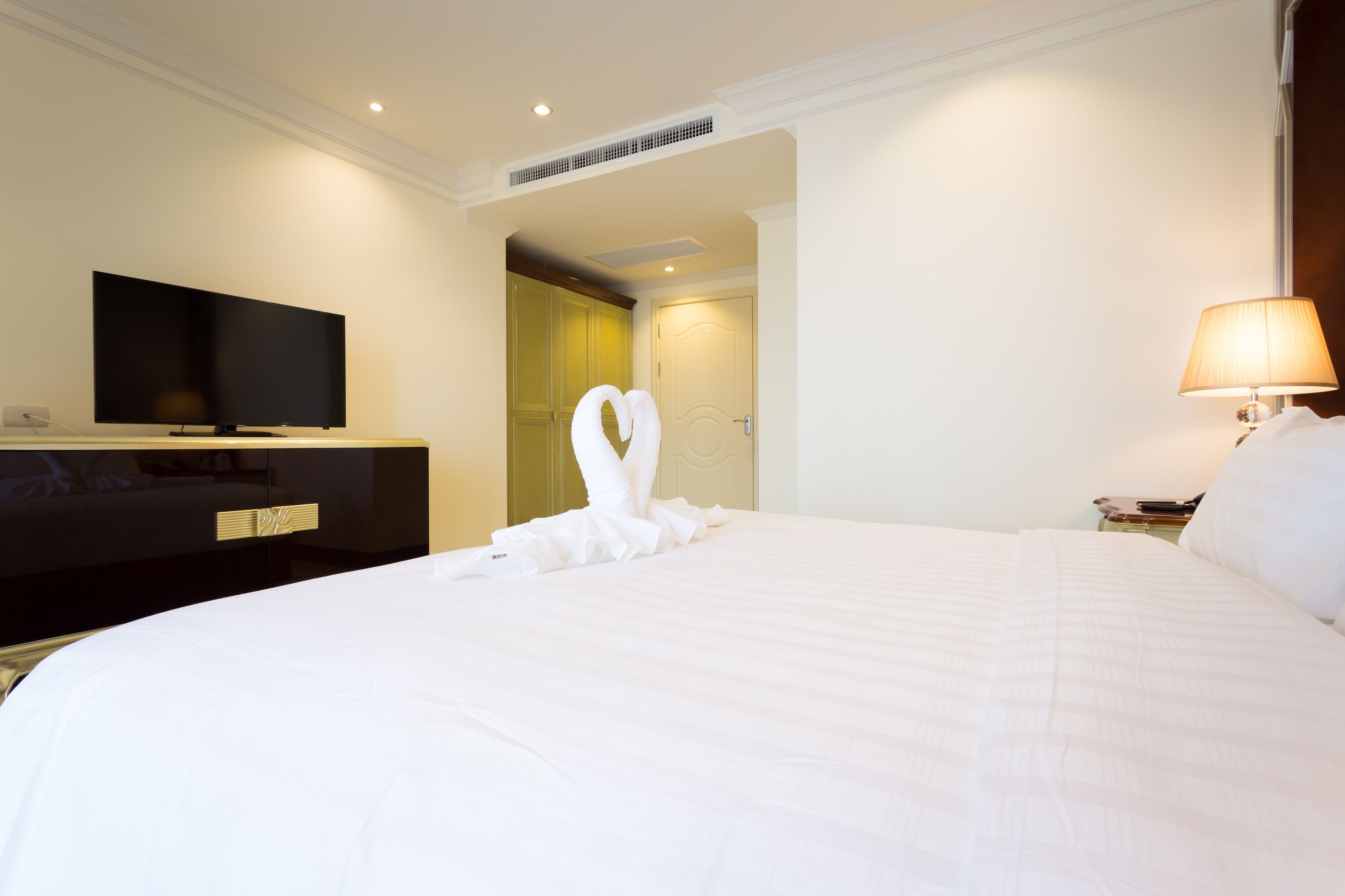 A hotel room with a large bed, a flat-screen TV on a stand, a bedside table with a lamp, and a wardrobe.