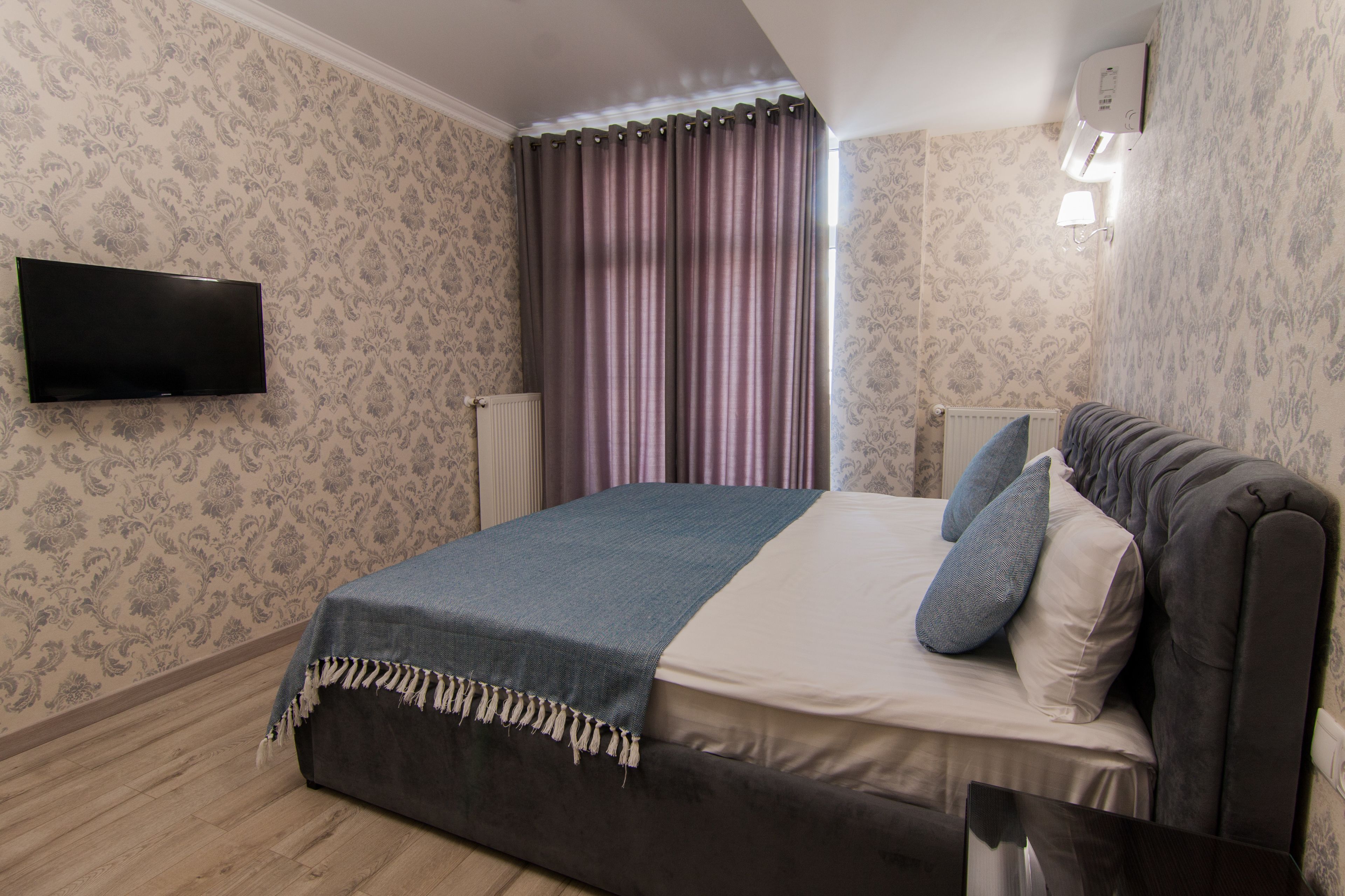 A bedroom with a bed, a flat-screen TV mounted on the wall, and a window with curtains.