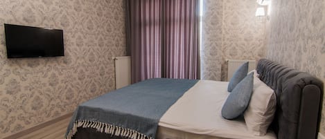 A bedroom with a bed, a flat-screen TV mounted on the wall, and a window with curtains.