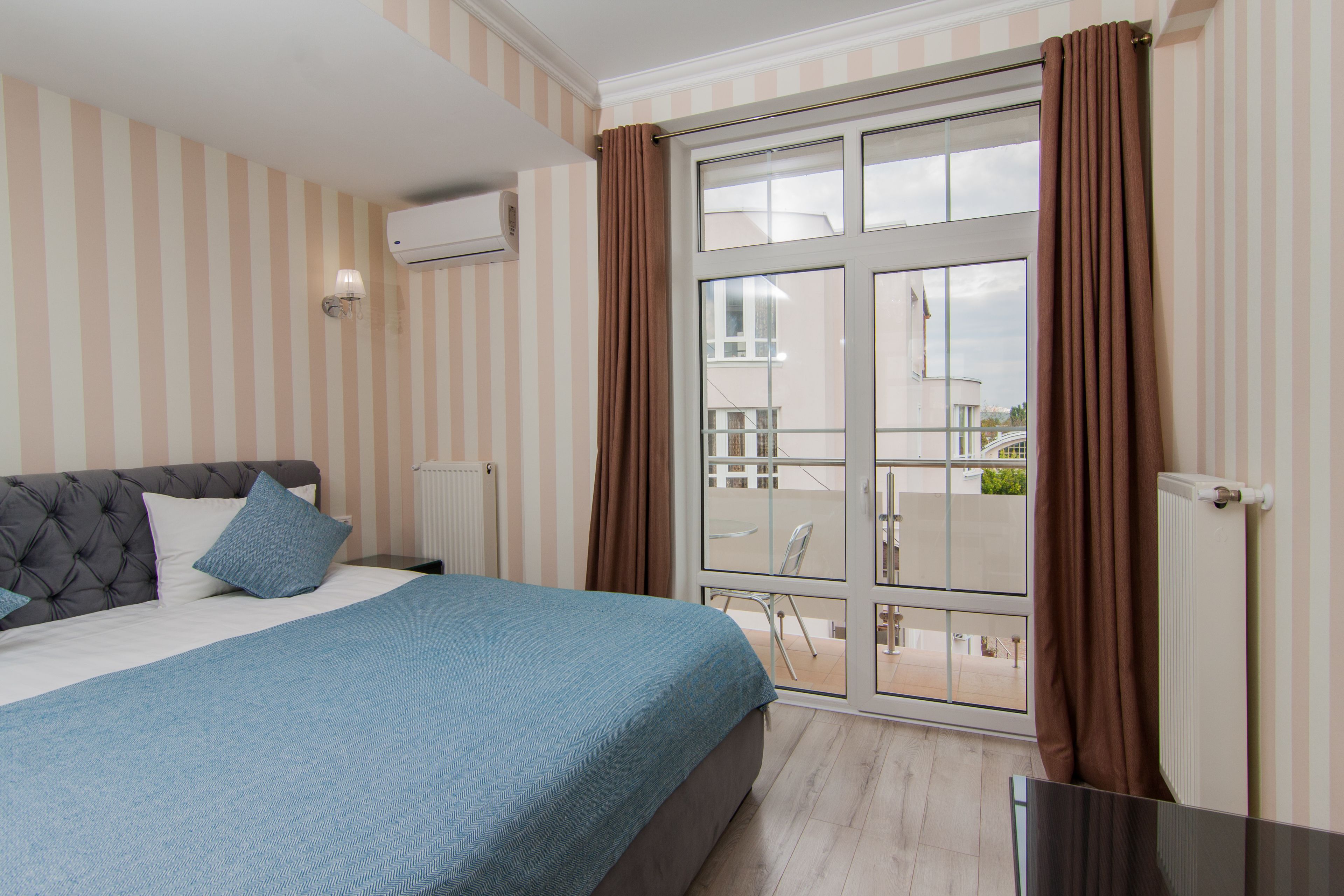 A bedroom with a bed, a window with curtains, a radiator, and a view of the outside balcony.