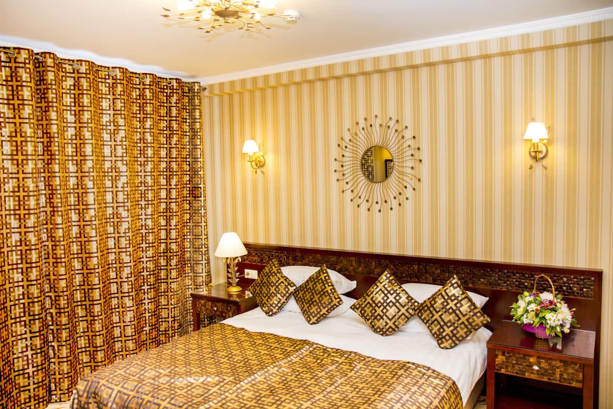 Une chambre d’hôtel avec un grand lit, des rideaux à motifs, un miroir décoratif, des appliques murales et une table de chevet agrémentée d’un arrangement floral.