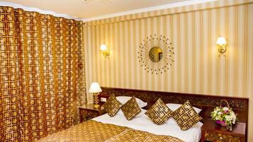 A hotel room with a large bed, patterned curtains, a decorative mirror, wall sconces, and a nightstand with a flower arrangement.