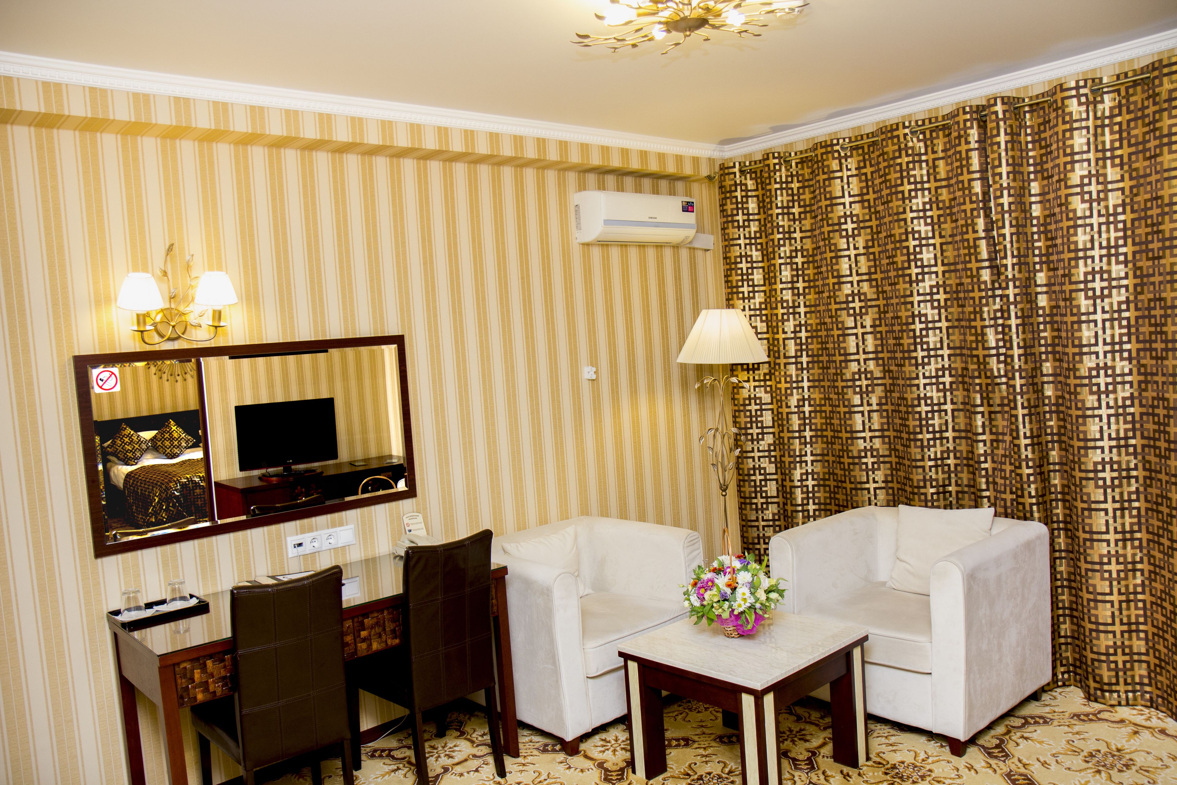 A hotel room with a striped wallpaper, a flat-screen TV, a desk with a chair, two sofas, a small table with a vase of flowers, and an air conditioning unit.