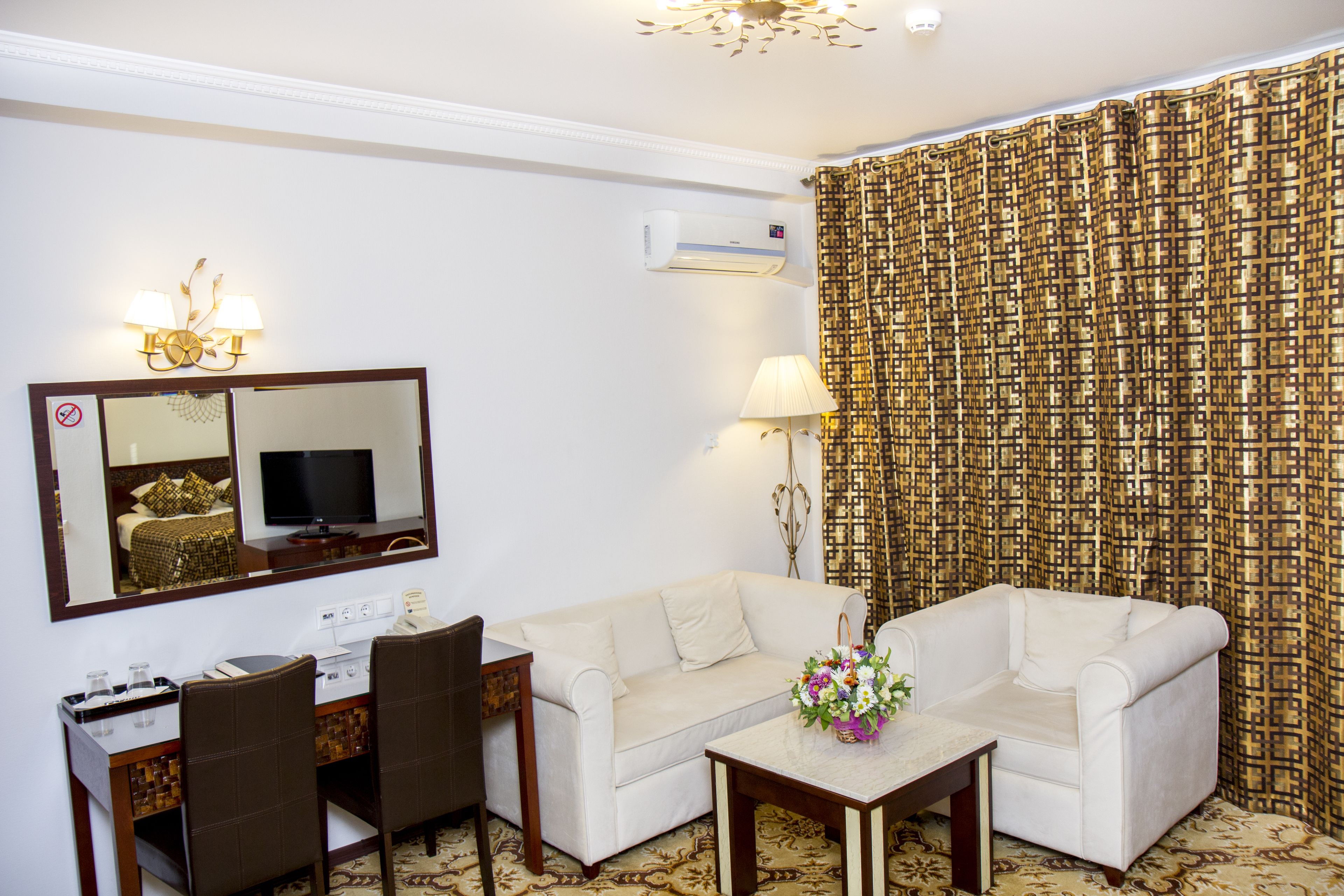 Habitación de hotel con sofá, dos sillones, una mesita con un jarrón de flores, un escritorio con computadora, televisor y aire acondicionado.
