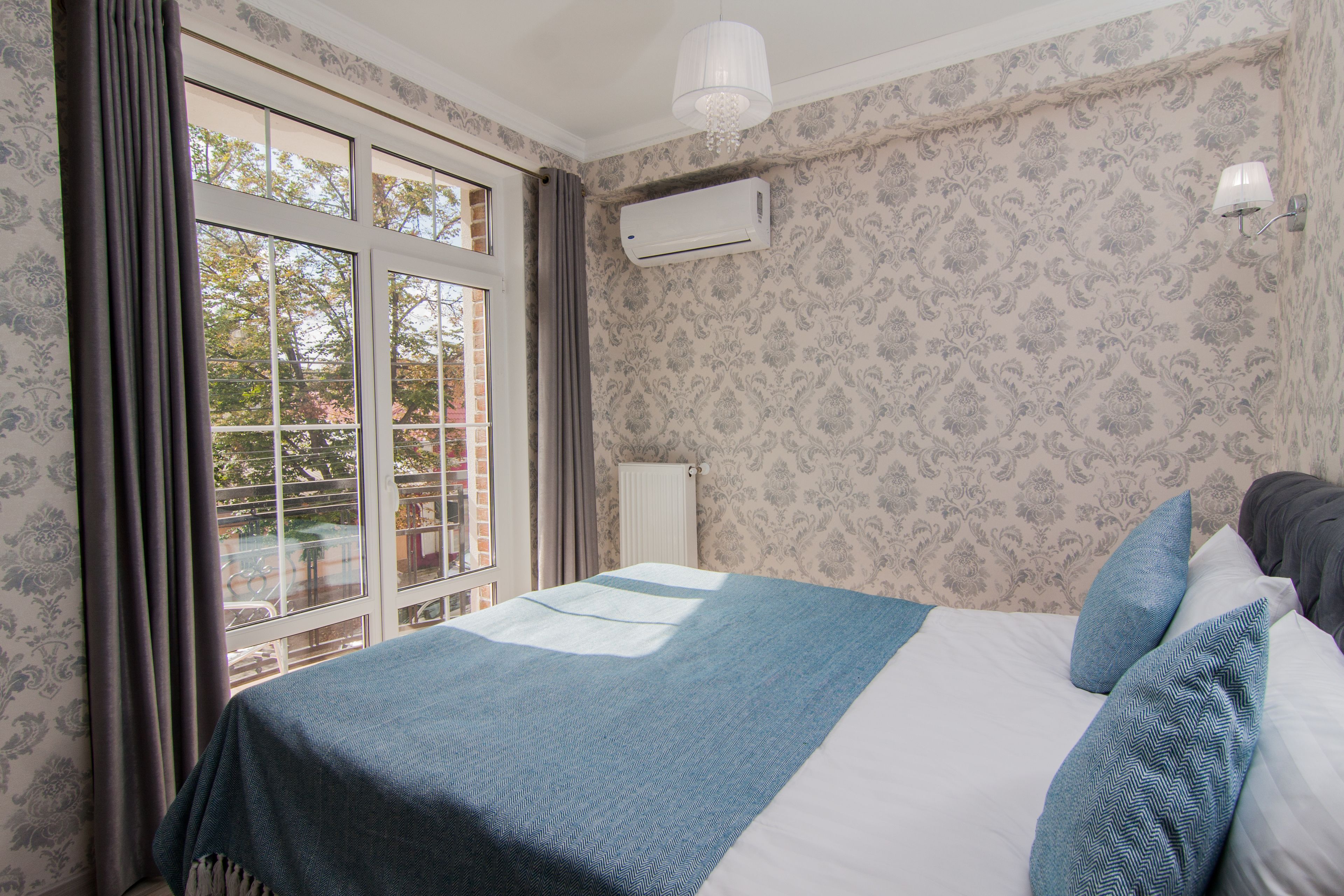 A bedroom with a large bed, a window with curtains, an air conditioning unit, and patterned wallpaper.