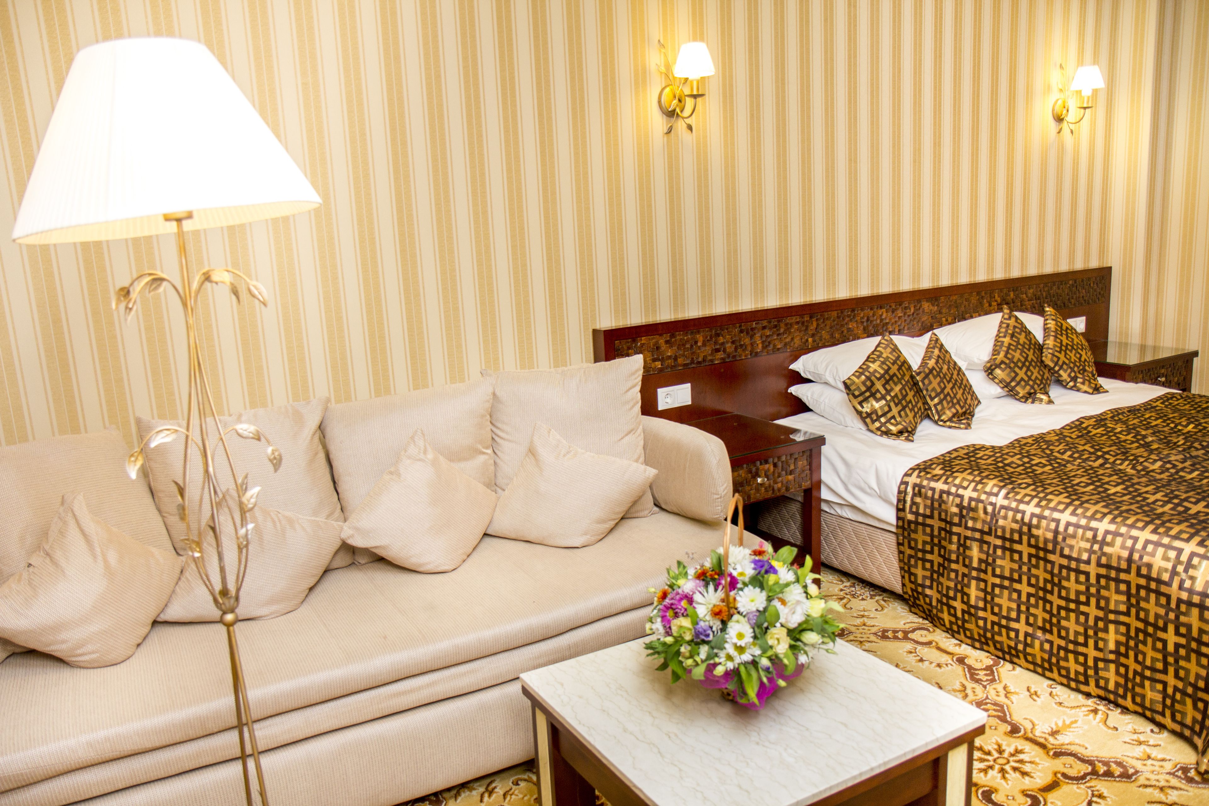 Hotelkamer met een bank, nachtkastje met bloemen, bed met behang en twee wandlampen.