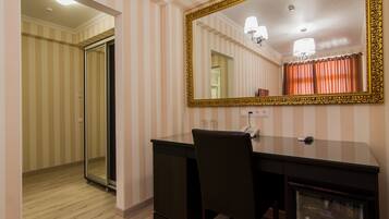 A hotel room with a striped wallpaper, a large mirror with an ornate frame, a desk with a chair, and a minibar.