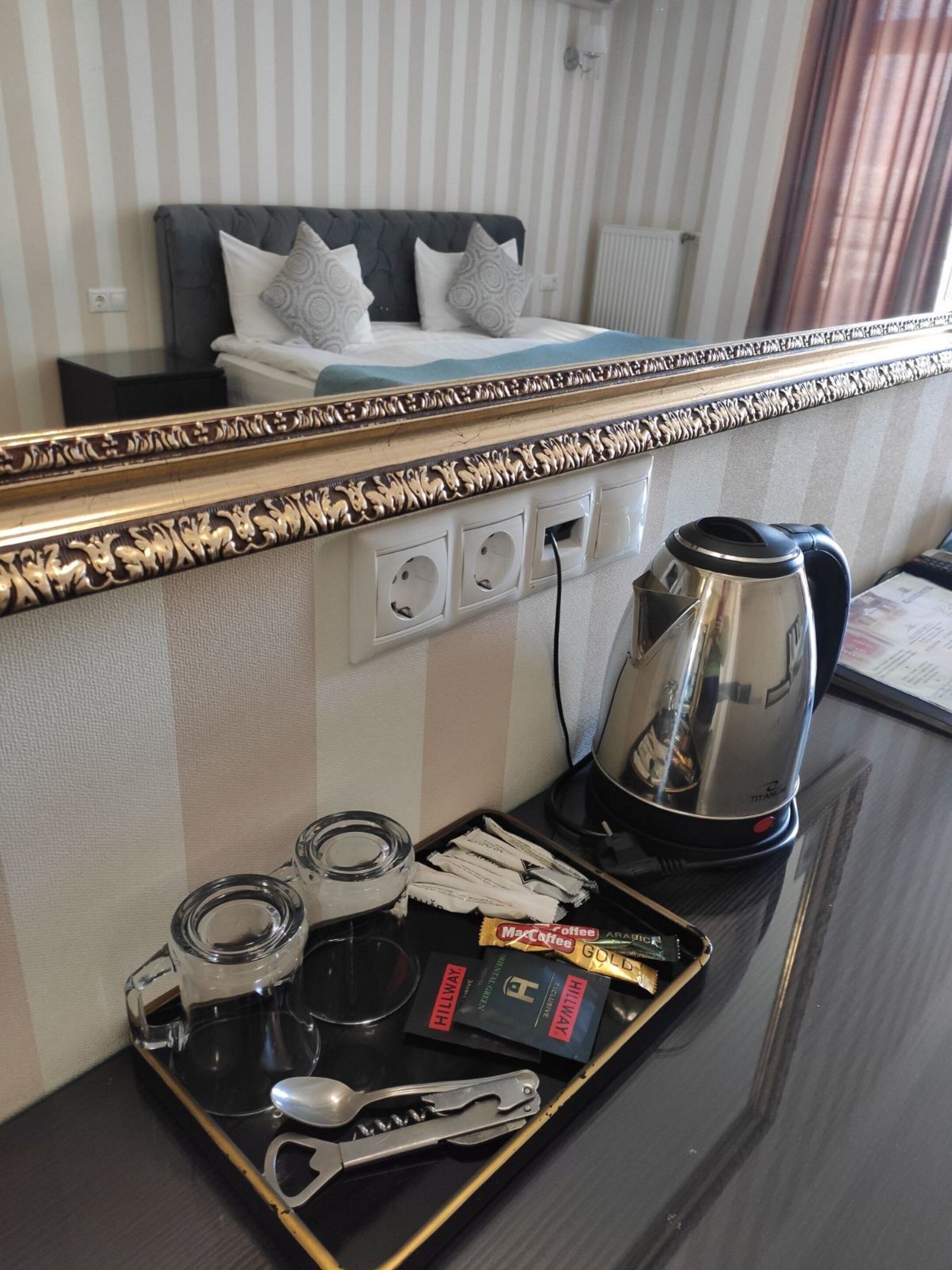 Habitación de hotel con cama, mesita de noche con tetera, bandeja con sobres de café y cuchara.
