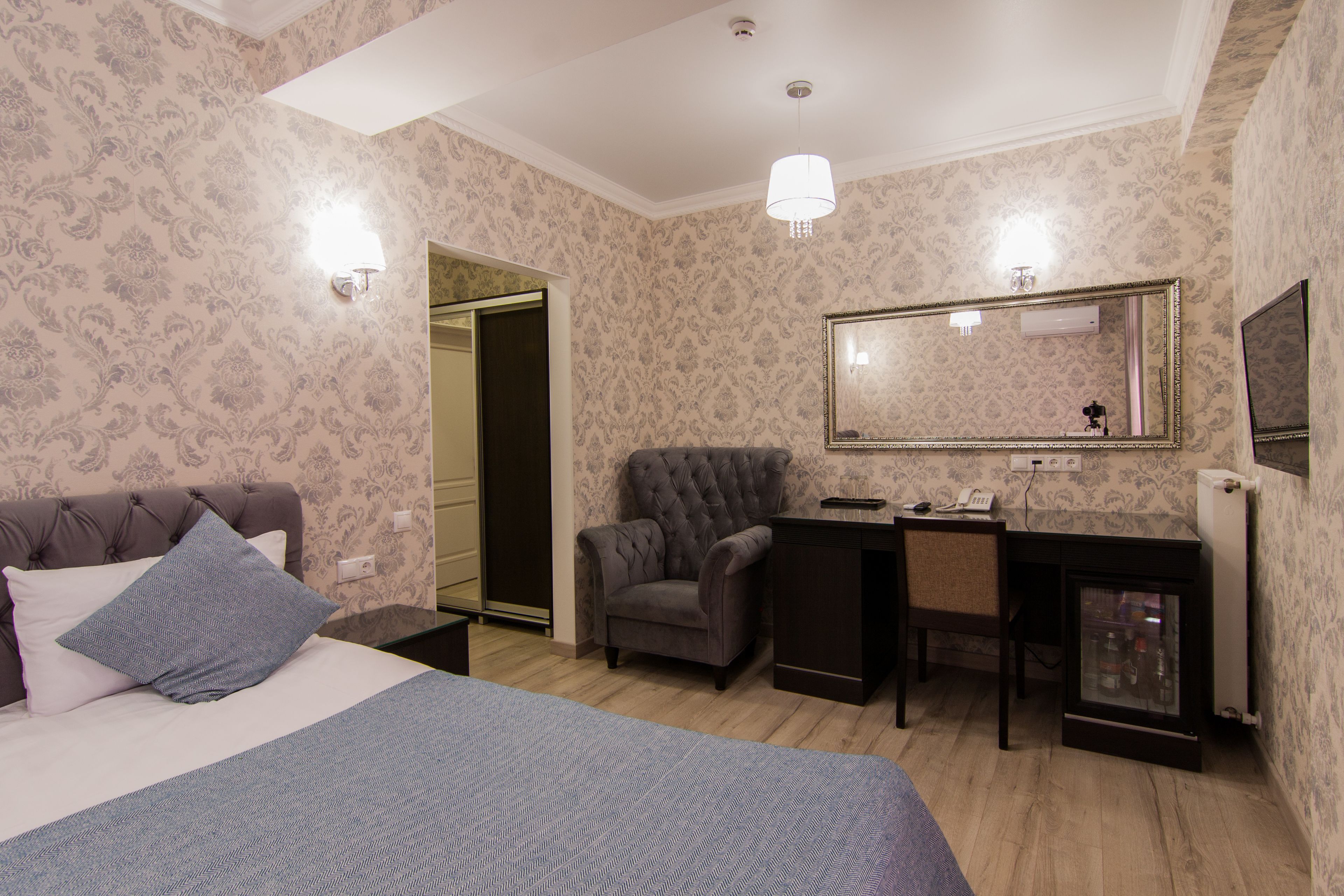 A bedroom with a bed, a desk, and a chair. There is a mirror on the wall and a door leading to another room.