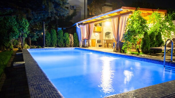 A swimming pool with a tiled edge, surrounded by a patio area with a pergola and trees. - Klassik Hotel (Chisinau)