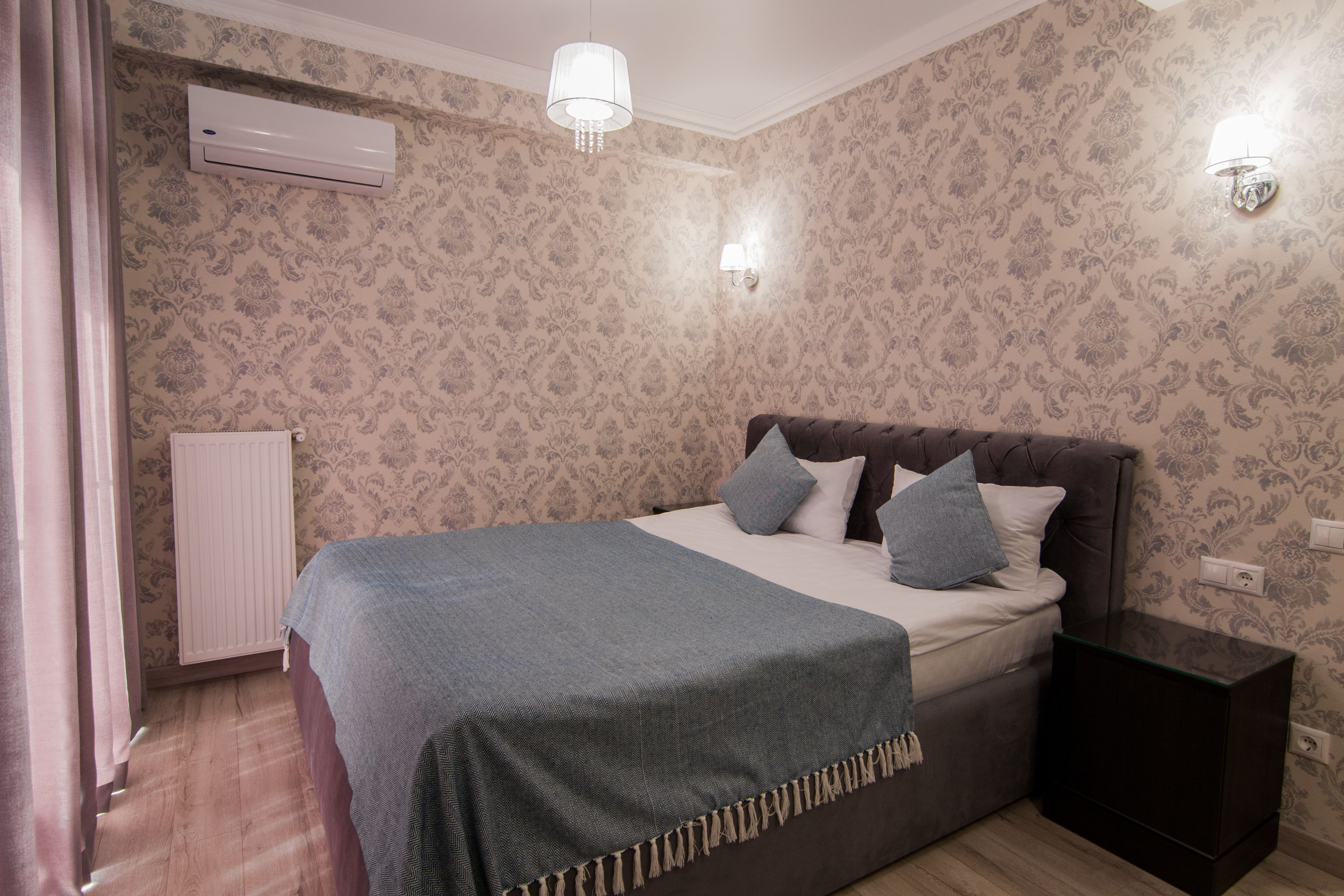 A bedroom with a bed, bedside table, wall-mounted light, air conditioning unit, and patterned wallpaper.