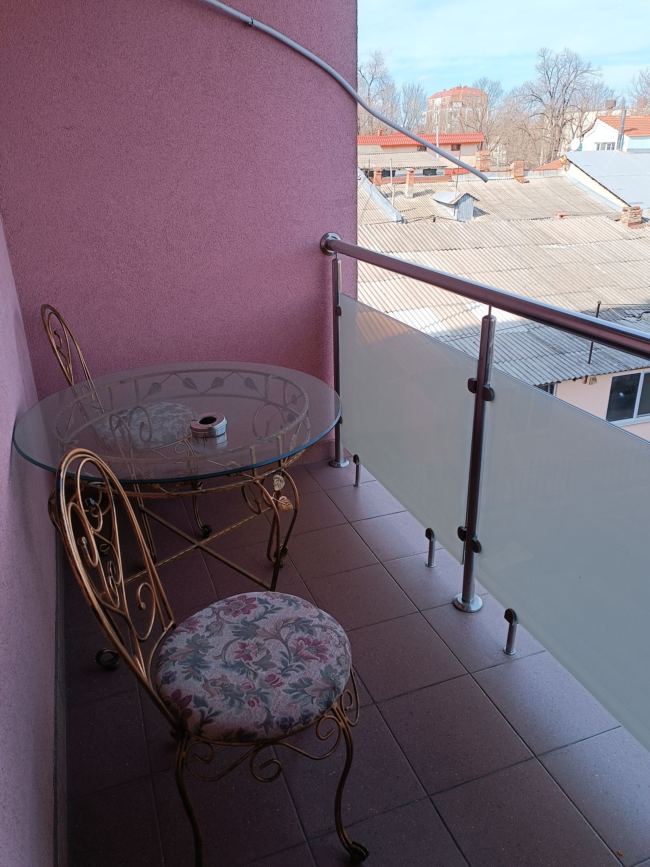 Un balcon avec une table en verre et des chaises en métal, un mur rose et une vue sur les immeubles.