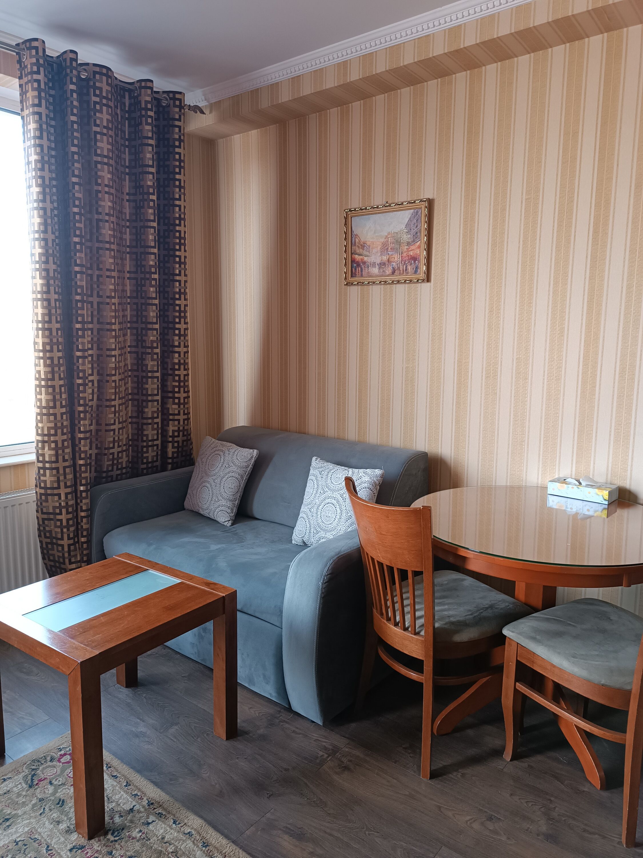 A cozy living room with a grey sofa, patterned cushions, a wooden coffee table, and a dining table with chairs. A framed picture hangs on the wall, and curtains are partially drawn.