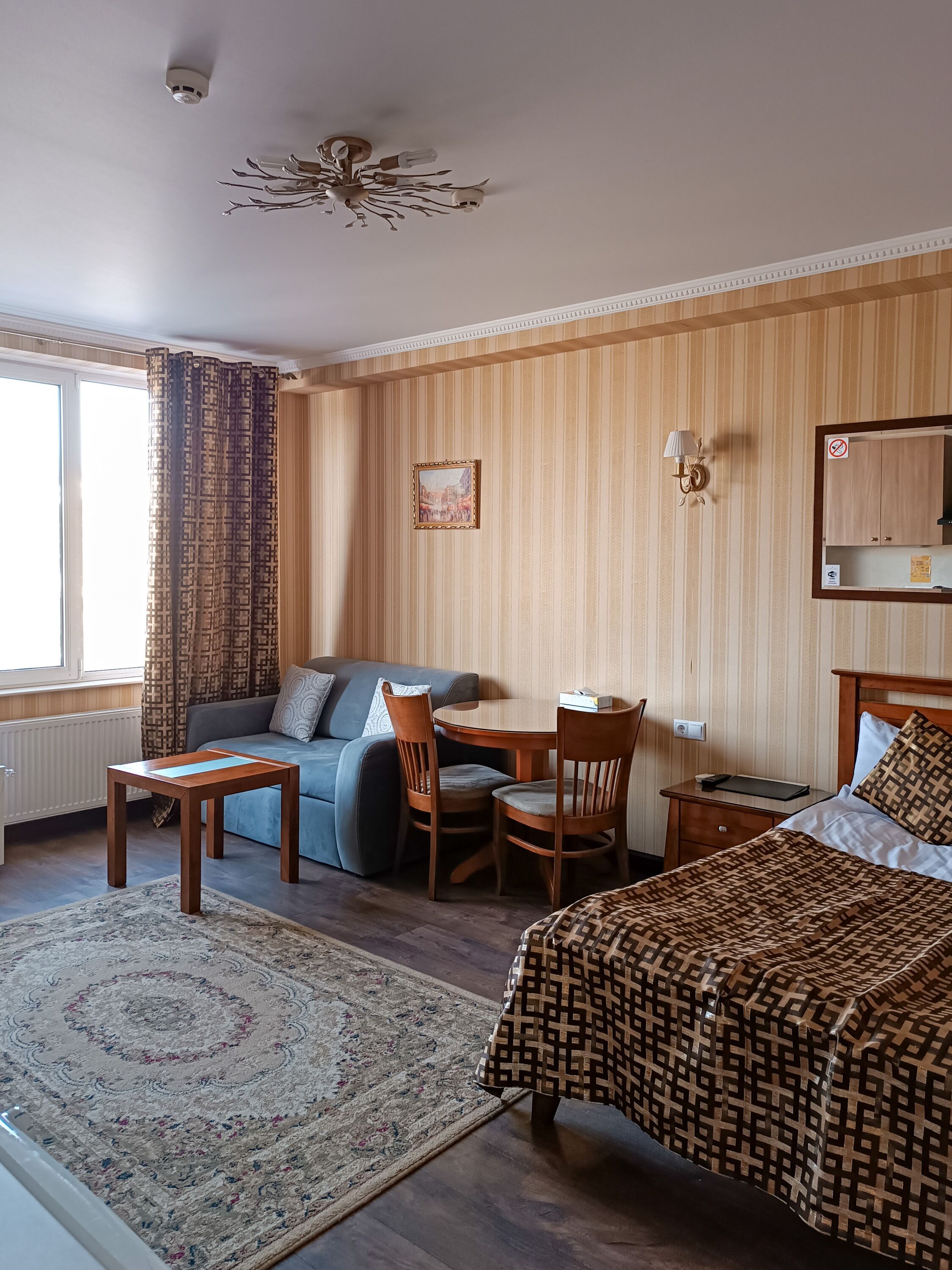 Quarto de hotel com cama, sofá, mesa de jantar com cadeiras, tapete pequeno, ventoinha no teto e janela com cortinas.