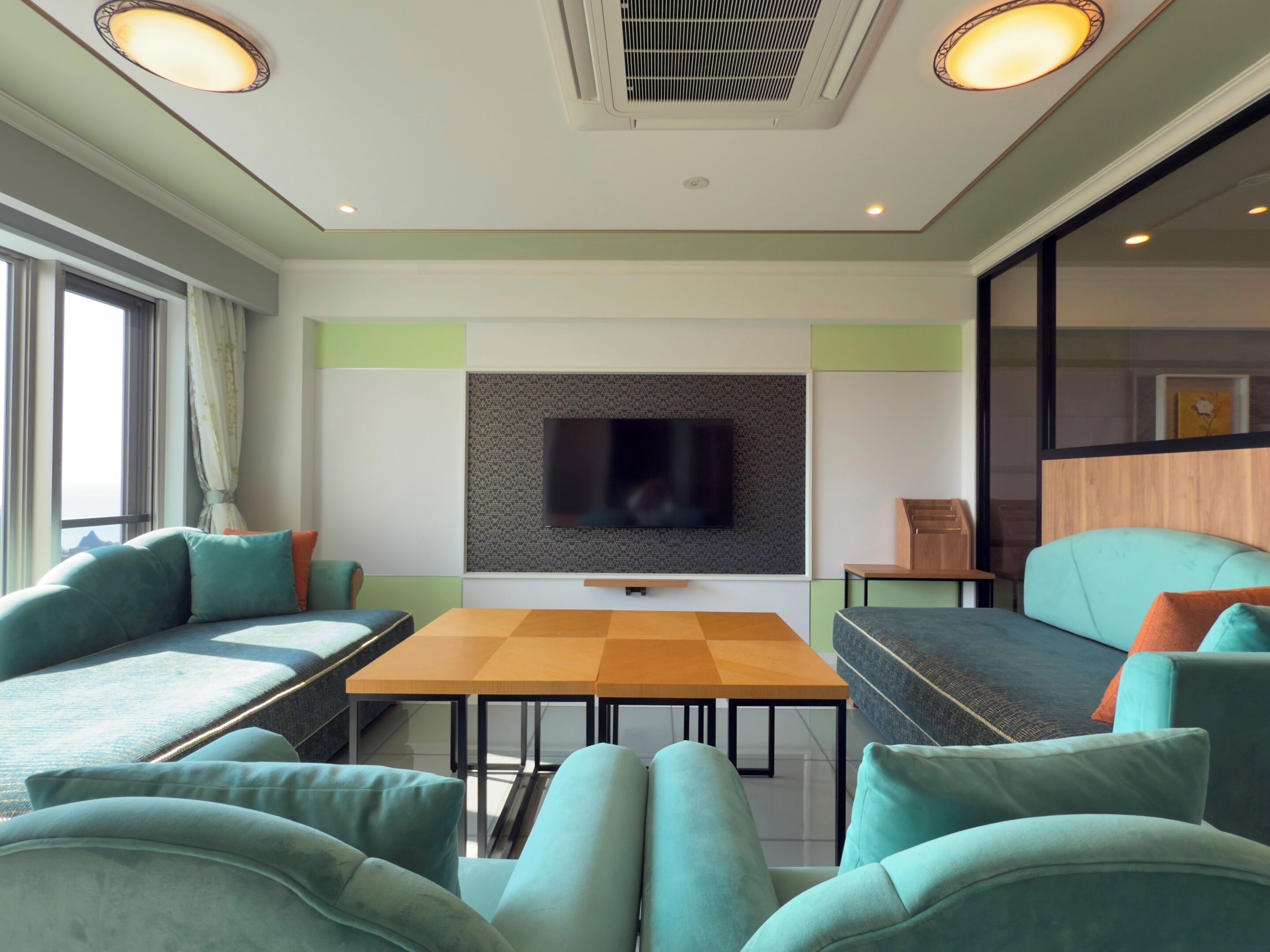 A modern living room with a flat-screen TV mounted on a patterned wall, a wooden coffee table, teal sofas, and a large window with curtains.