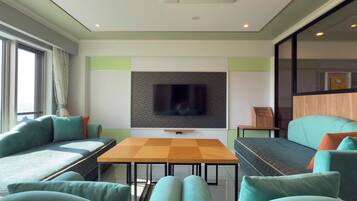 A modern living room with a flat-screen TV mounted on a patterned wall, a wooden coffee table, teal sofas, and a large window with curtains.