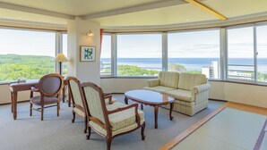 Ein geräumiger Raum mit großem Fenster und Blick auf den Wald und das Meer. Zwei Sofas, ein Tisch und zwei Stühle sind vorhanden.