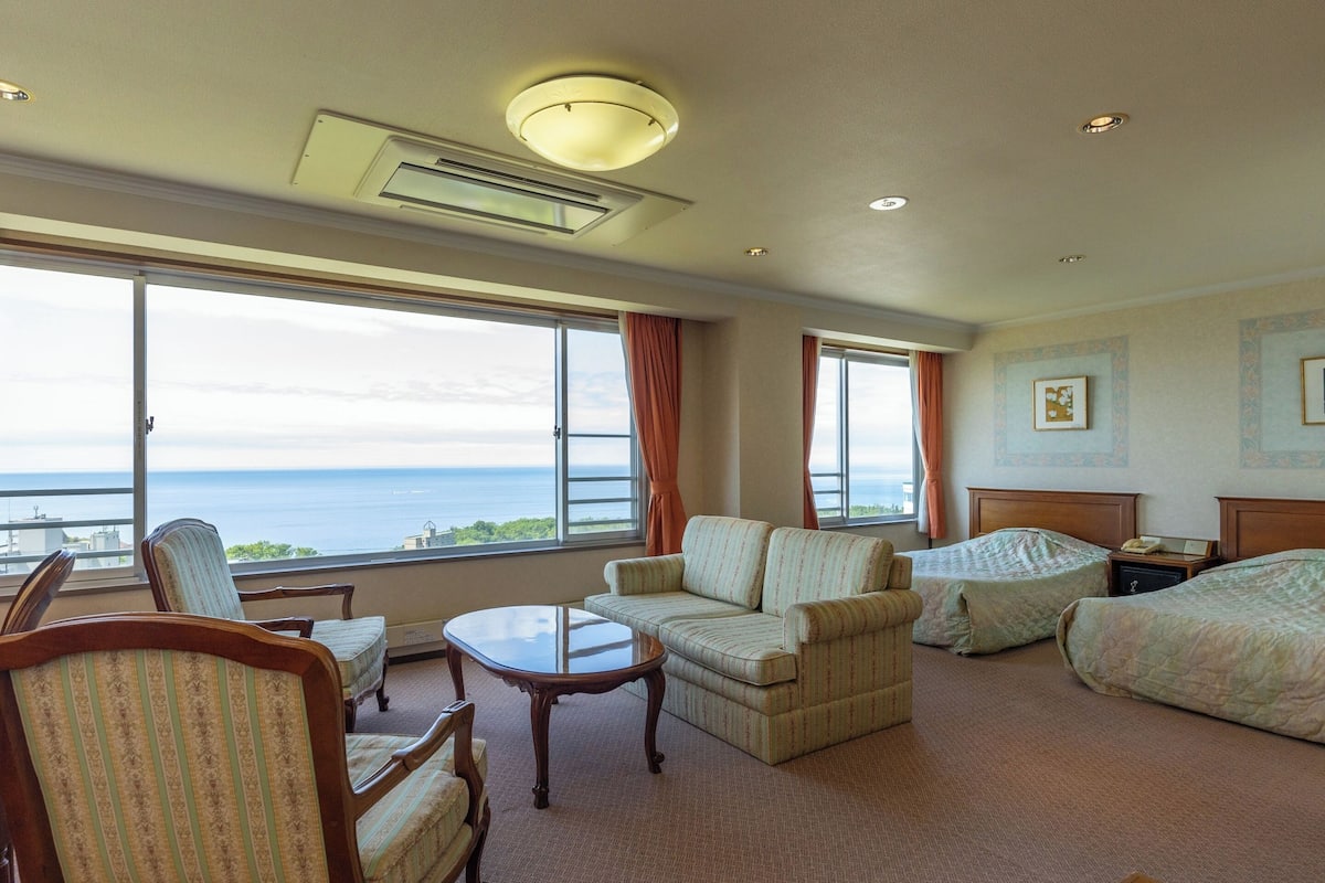 Un dormitorio de hotel con un ventanal que ofrece vistas al mar, un conjunto de sofás, una mesita y una cama.