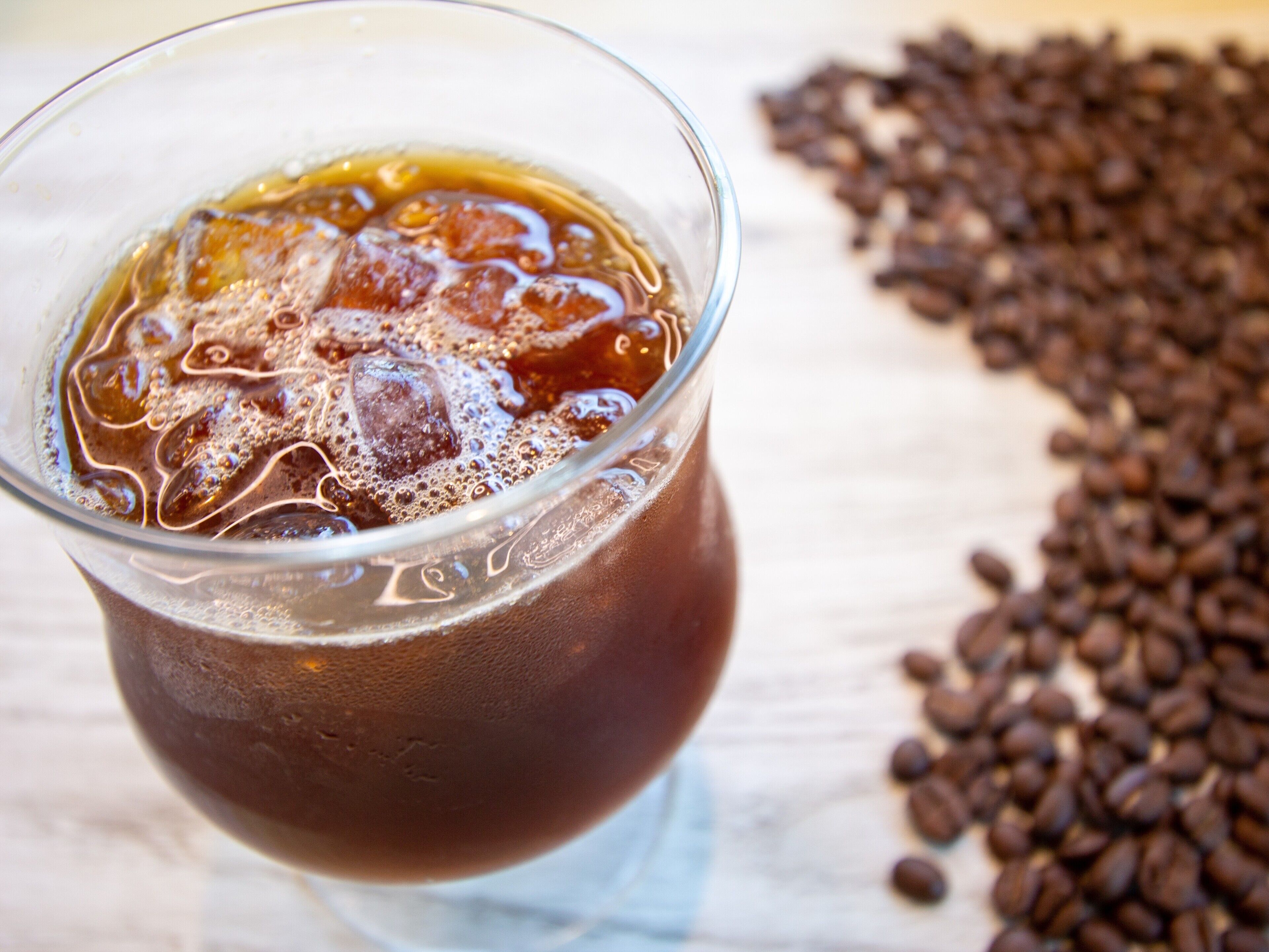 Un vaso de café helado con cubitos sobre una superficie de madera, rodeado de granos de café.