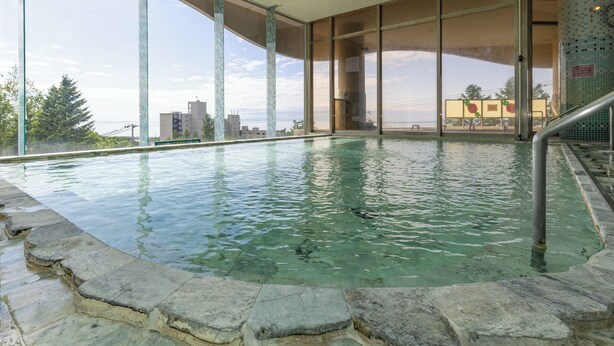 Ein Innenpool mit klarem Wasser, Steinstufen zum Ein- und Ausstieg, große Fenster mit Blick auf Gebäude und Bäume sowie eine geflieste Wand mit einer Uhr.