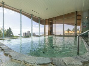 Una piscina interior con agua cristalina, escalones de piedra que bajan a la piscina, ventanas amplias con vistas a edificios y árboles, y una pared de azulejos con un reloj.