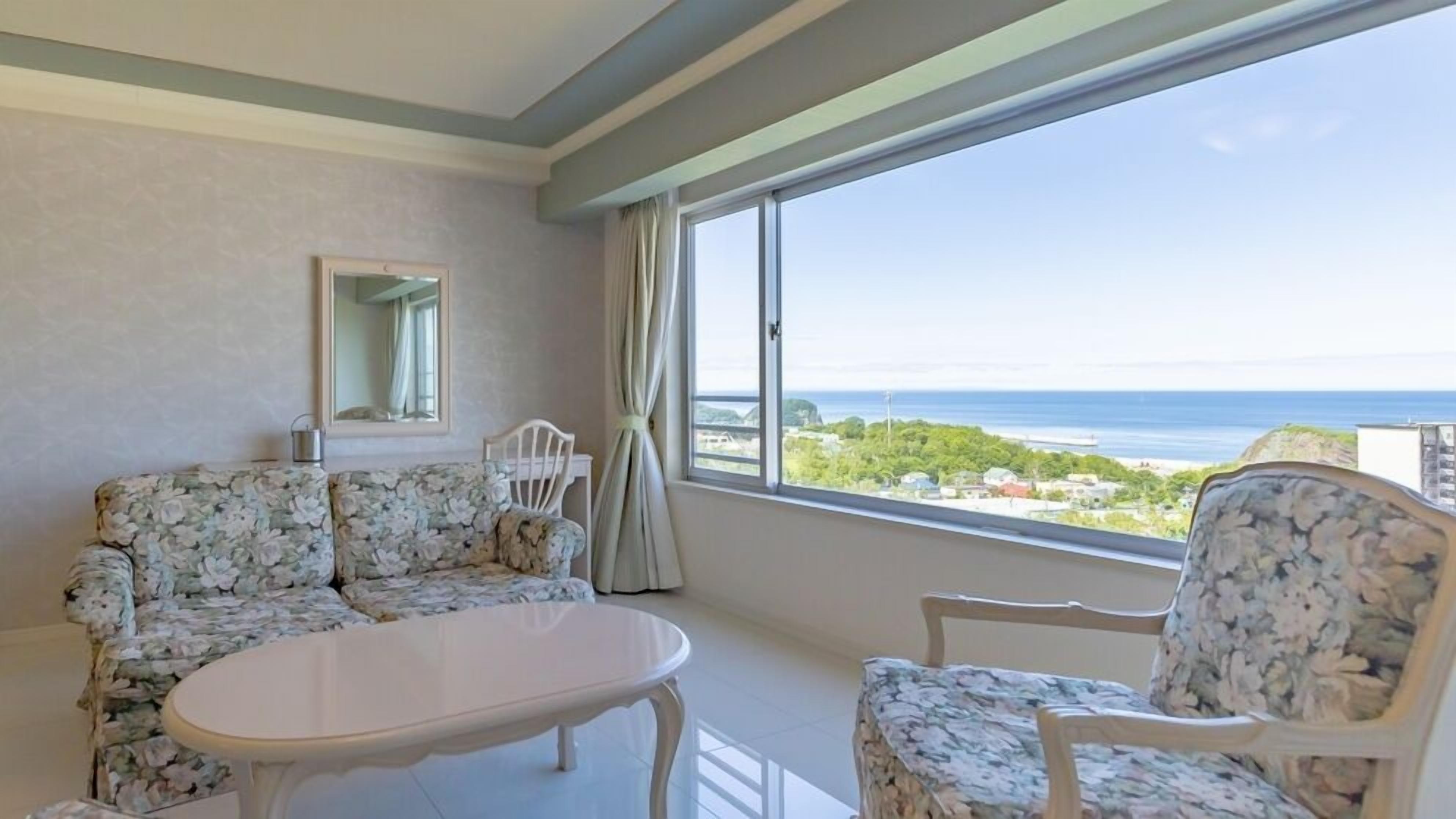 A room with a sofa, a chair, a small table, a mirror, and a large window with a view of the sea.