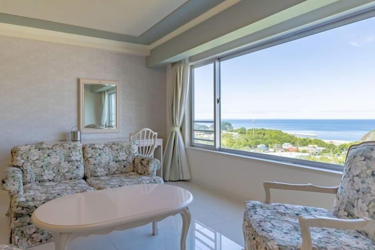 Una habitación con sofá, una silla, una mesita, un espejo y un ventanal con vistas al mar.