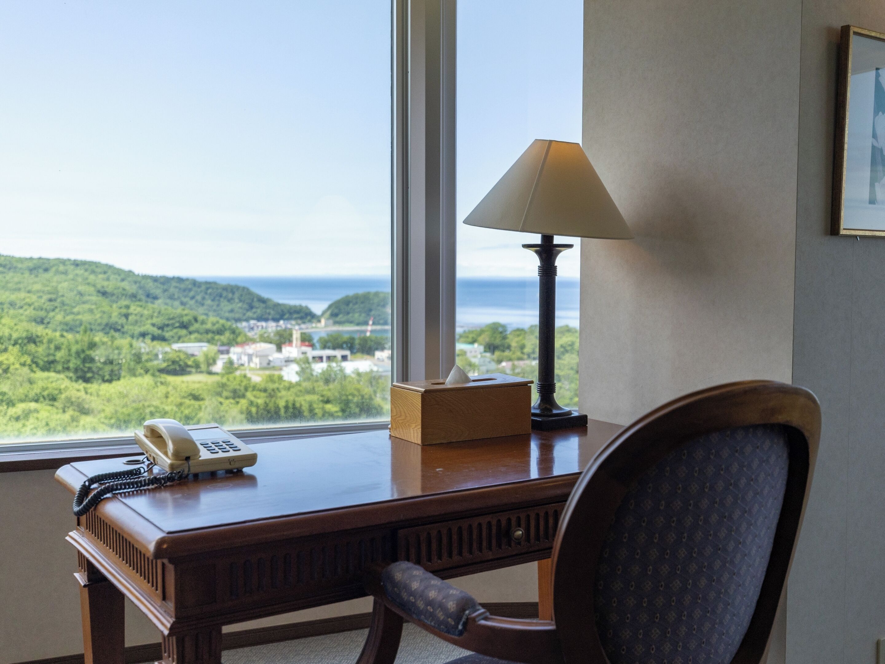 A hotel room with a desk, a telephone, a lamp, and a chair. The room has a large window with a view of a green landscape and a body of water.
