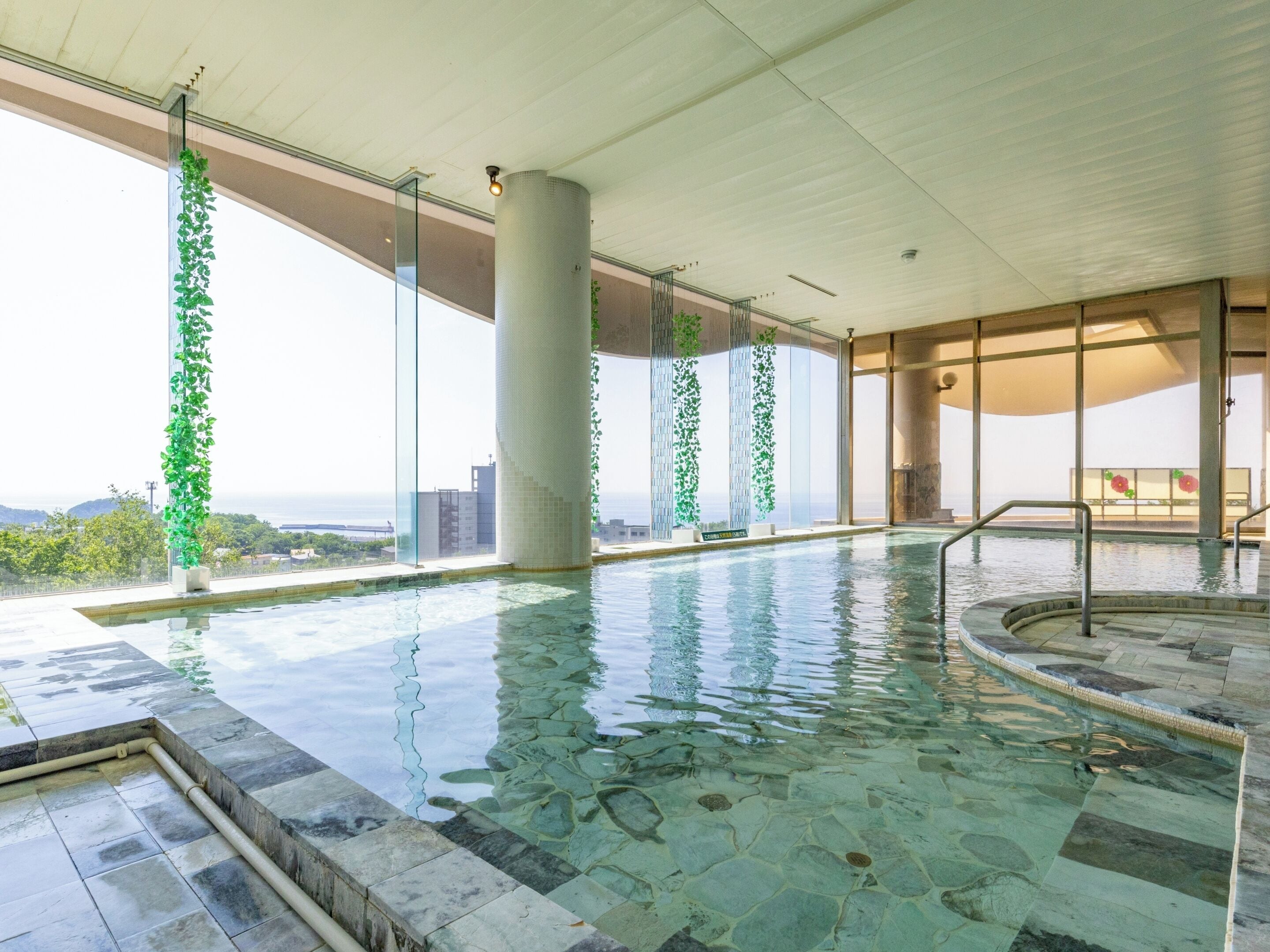 An indoor swimming pool with clear water, stone flooring, and green plants on columns.