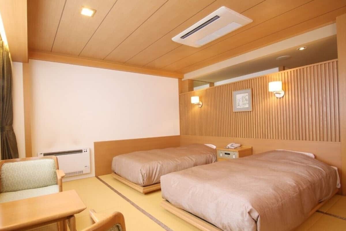 Habitación de hotel con dos camas, un escritorio y una silla. Hay un calentador instalado en la pared y una mesita de noche con una lámpara.