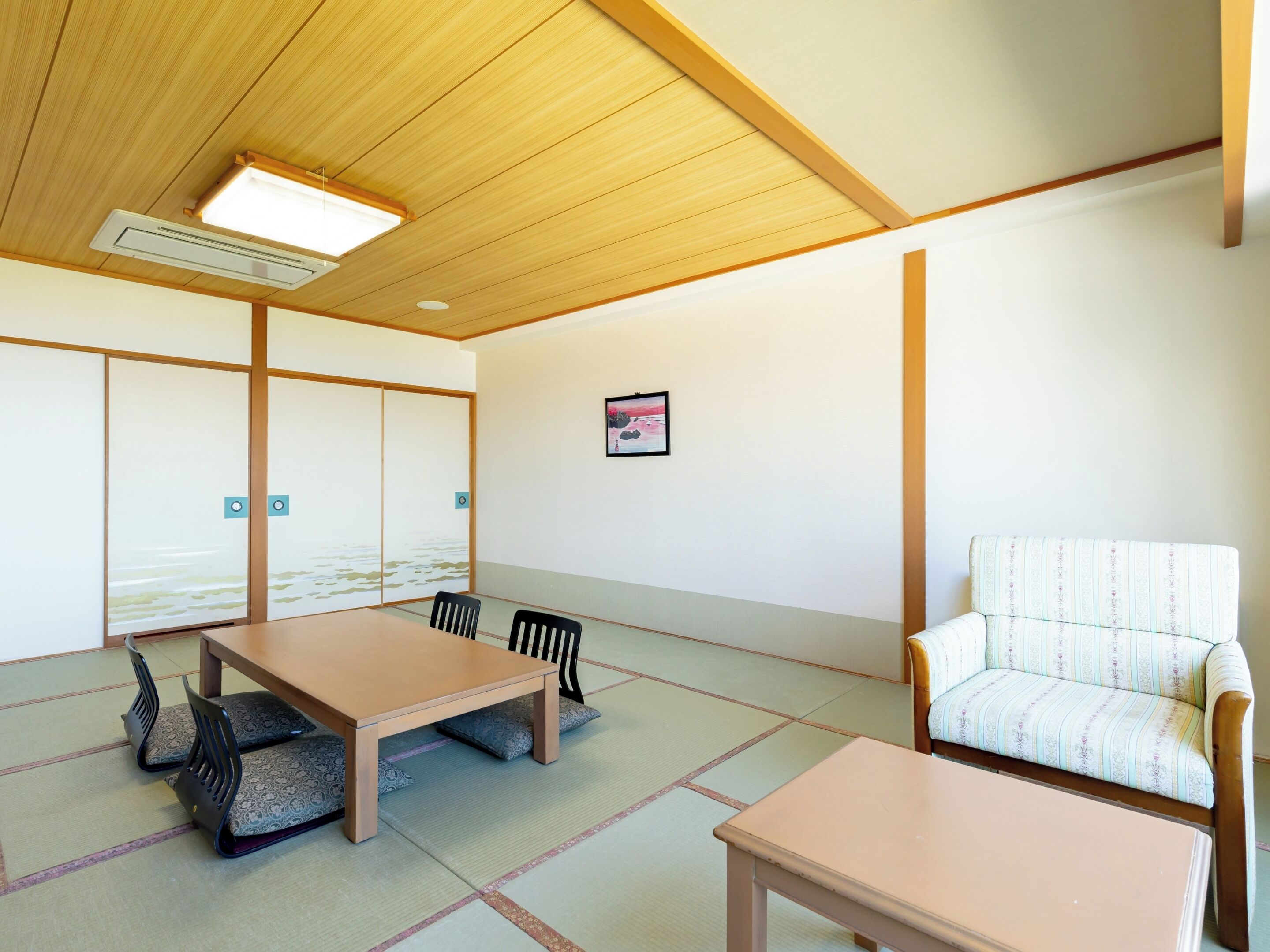 Une pièce dotée d’un sol recouvert de tatamis, d’une table en bois, de chaises, d’un canapé, d’un ventilateur de plafond et d’un climatiseur fixé au mur.