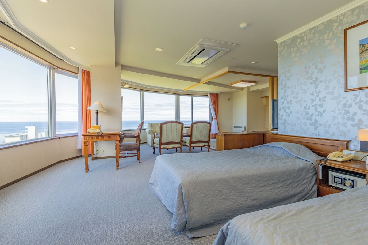 Habitación de hotel con dos camas, un escritorio, una silla y un amplio ventanal con vistas al océano.