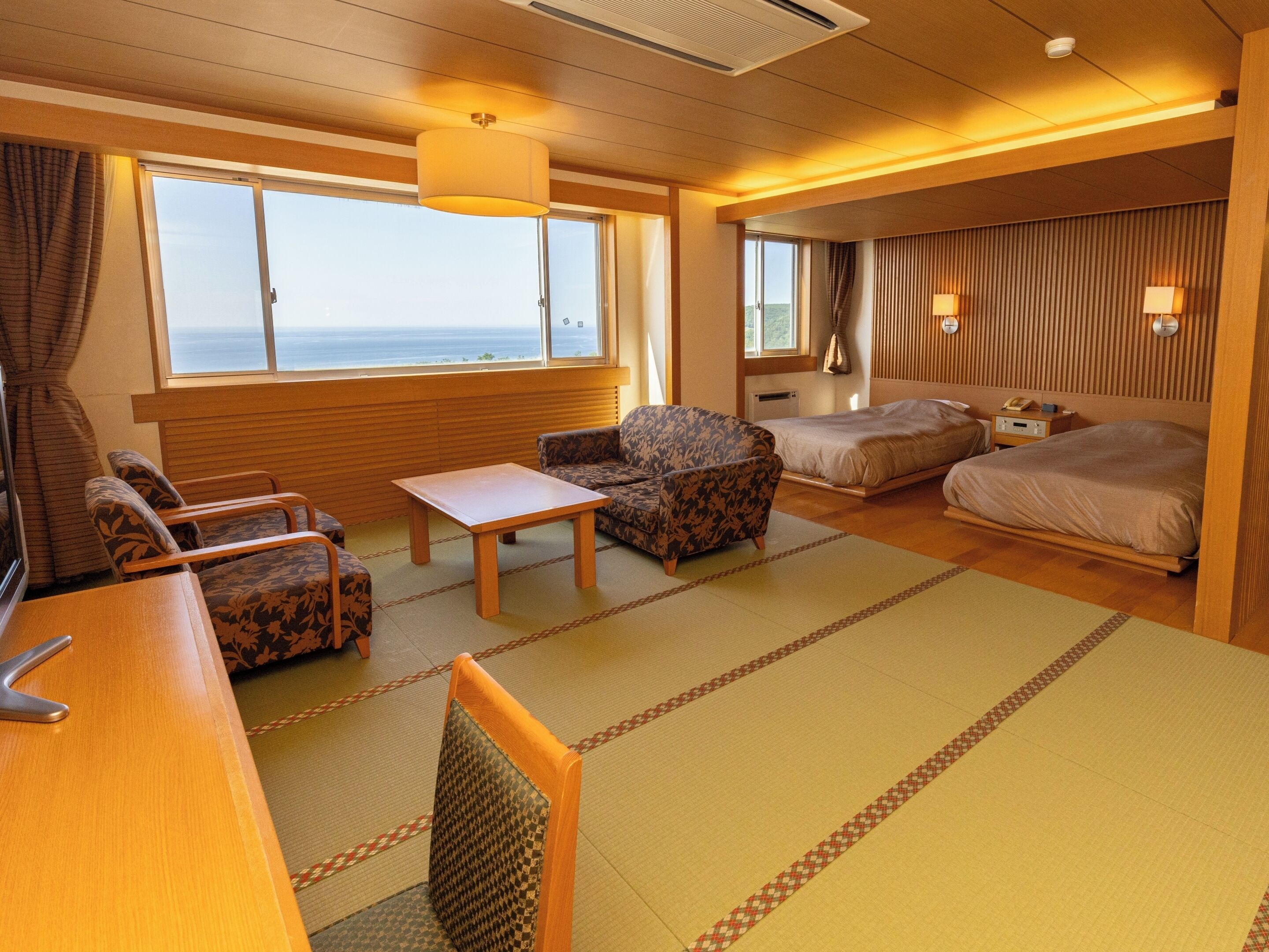 A room with a bed, a sofa, a chair, a table, and a view of the sea through large windows.